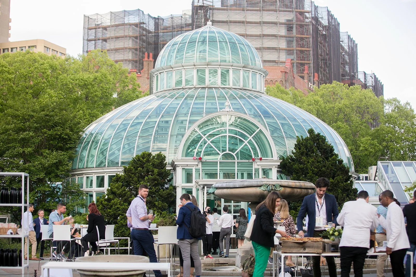 A glass greenhouse with a domed roof amid green trees, with people at outdoor tables and a buffet in the foreground