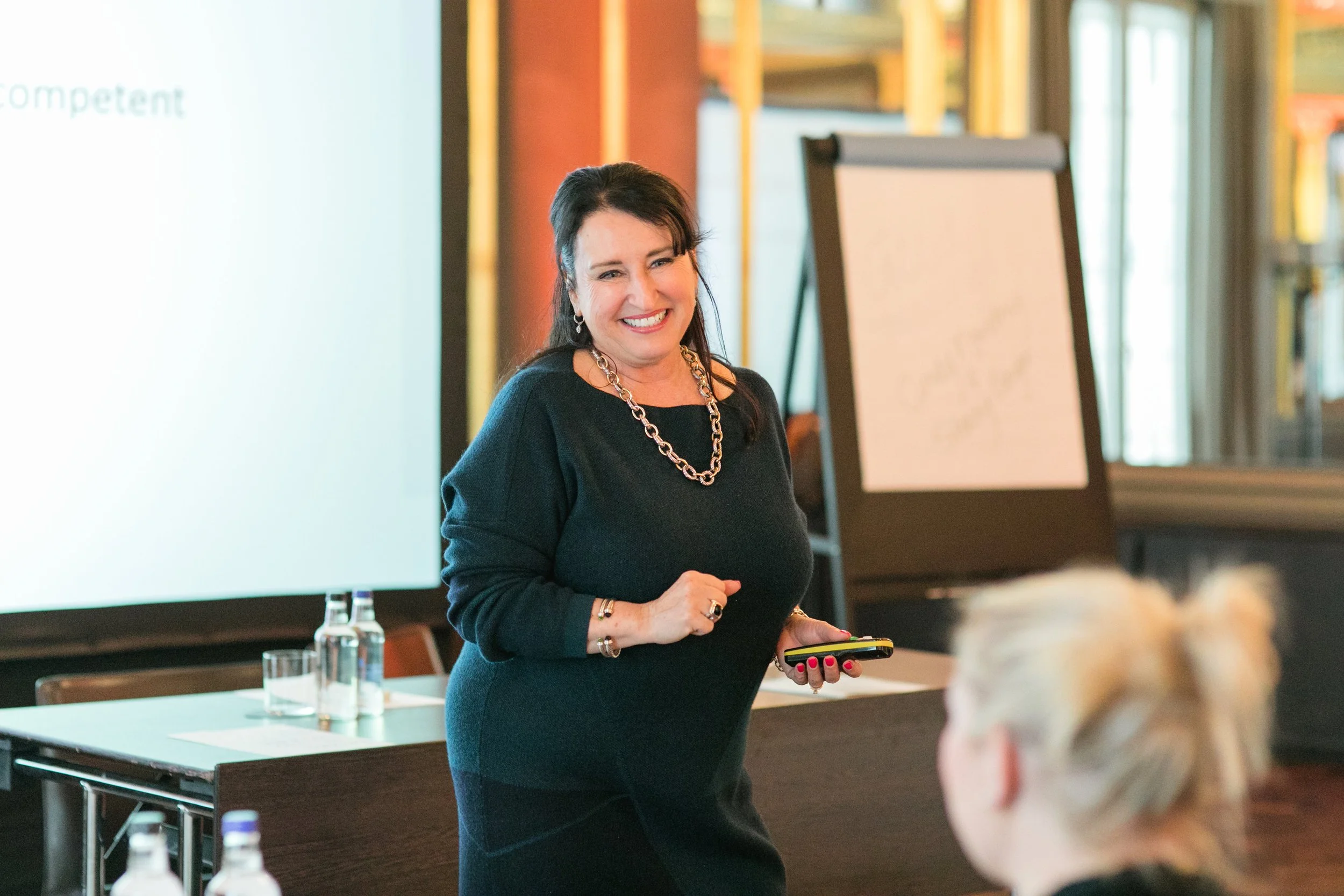 A woman smiling while speaking at a presentation in a conference room. She is wearing a black top with a chunky necklace and holding a remote control. There is a whiteboard behind her and some tables with water bottles and paper in the foreground.
