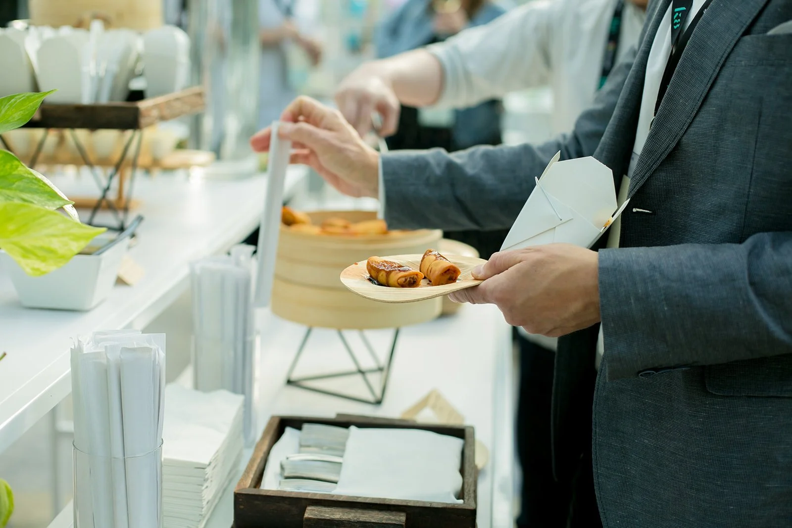 People at a buffet table serving food, including spring rolls and other appetizers, with disposable chopsticks and utensils.