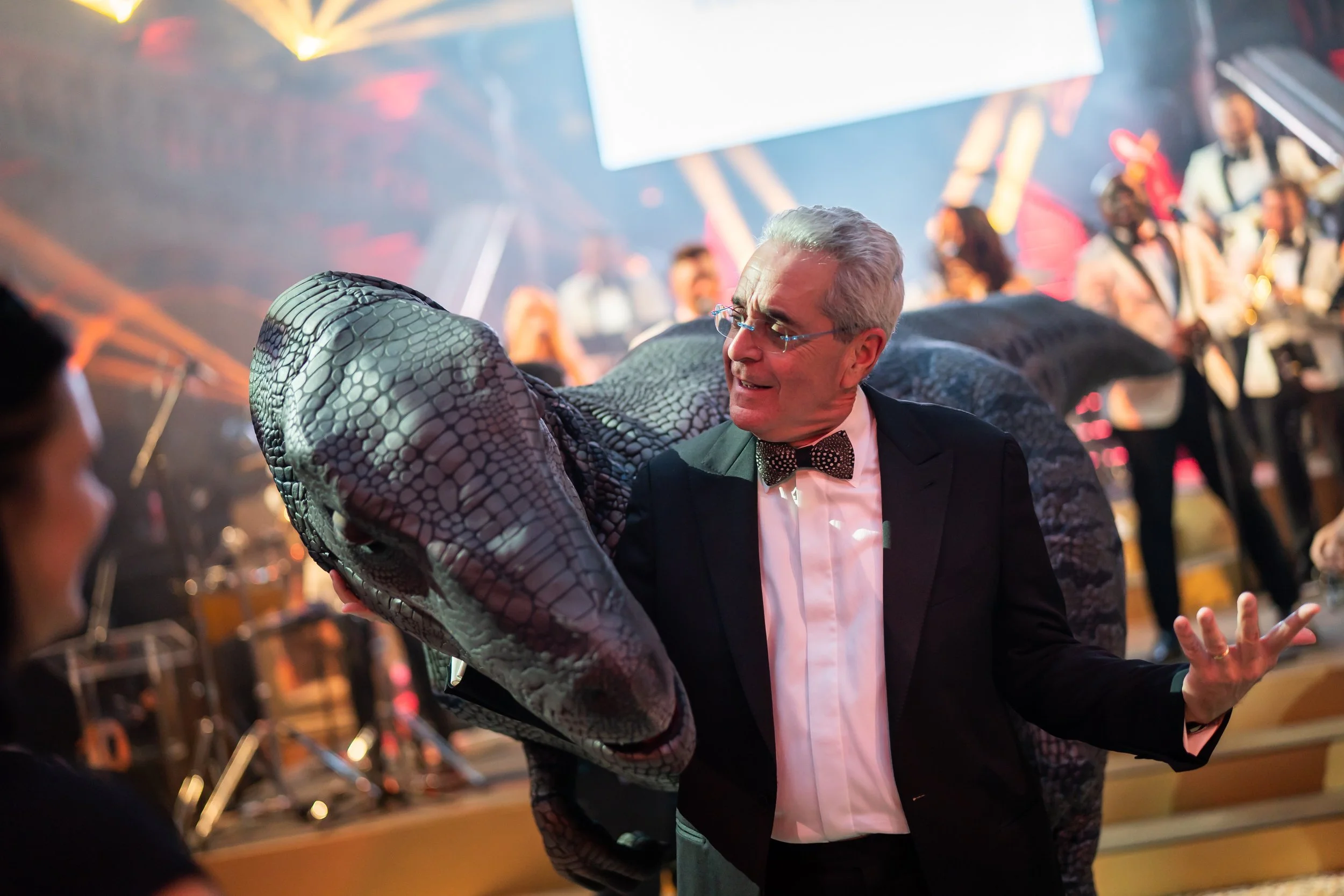 A man in a tuxedo and bowtie dancing with fake dinosaur at The Natural History Museum at a lively event with colorful lights and people in the background.