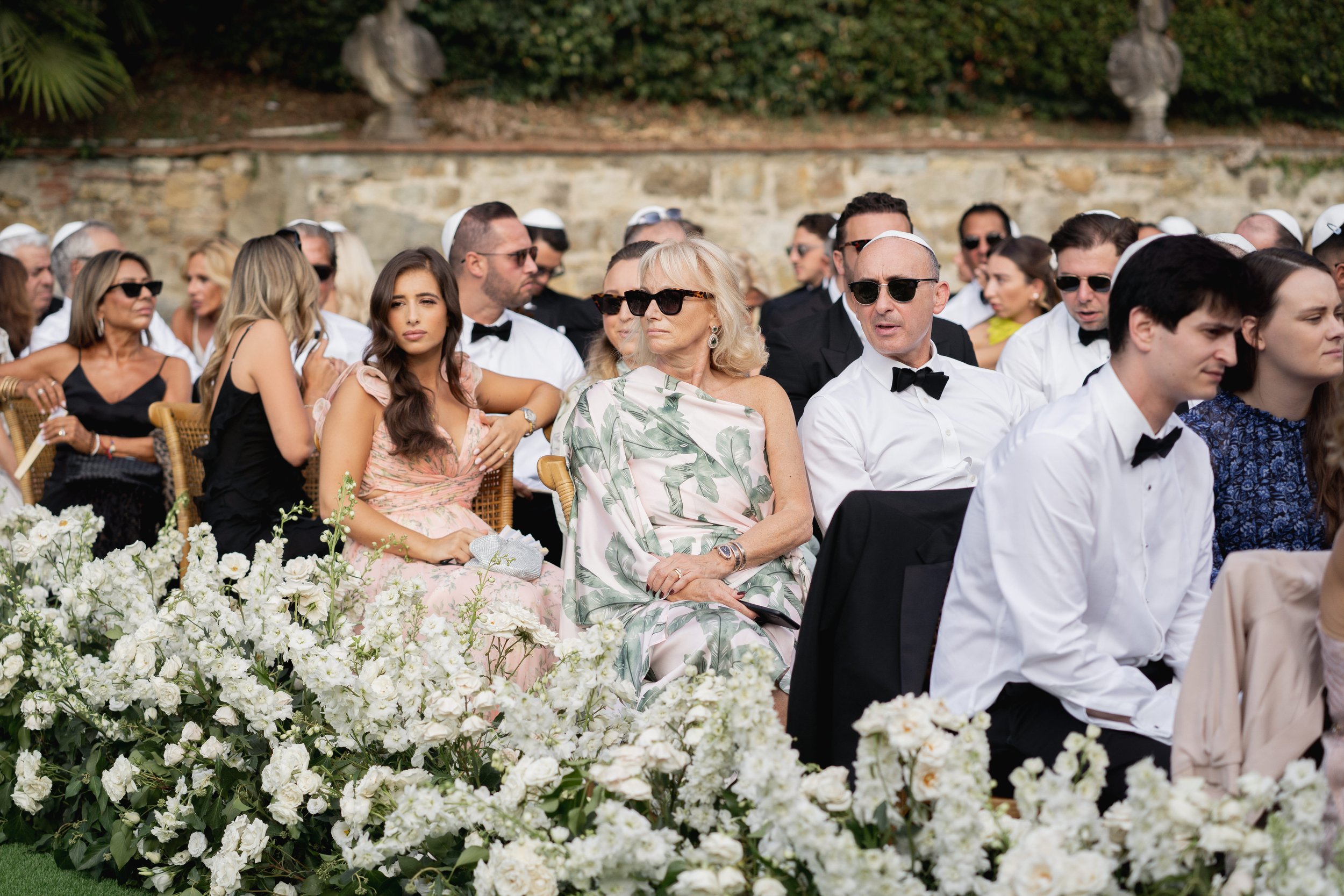 People dressed in formal attire, sitting outdoors near a stone wall, attending a luxury jewish wedding , with white flowers in the foreground ar Villa Palmieri