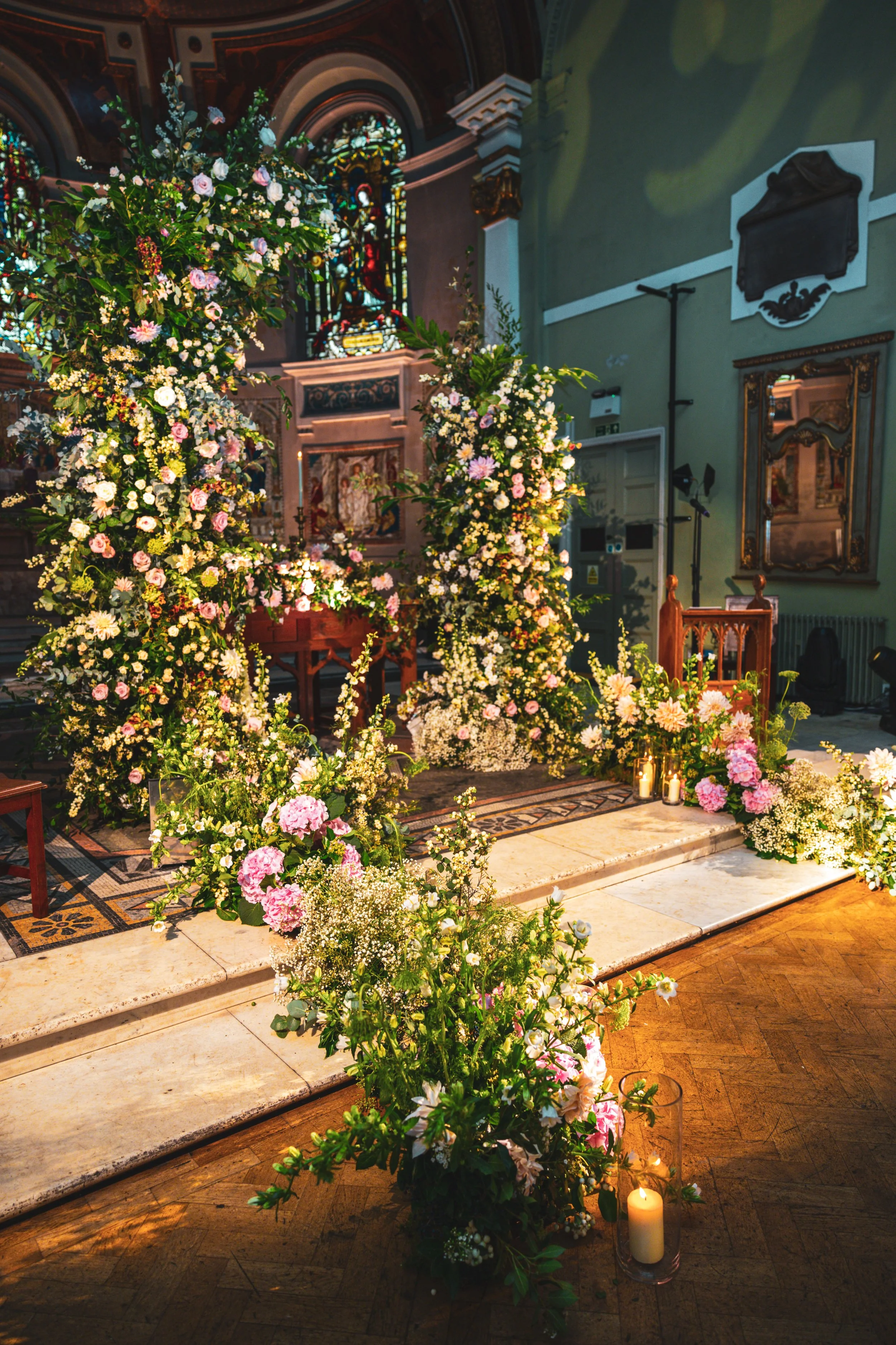 Interior of a church decorated for a wedding, with an altar surrounded by numerous pink and white flower arrangements, candles, and stained glass windows in the background.