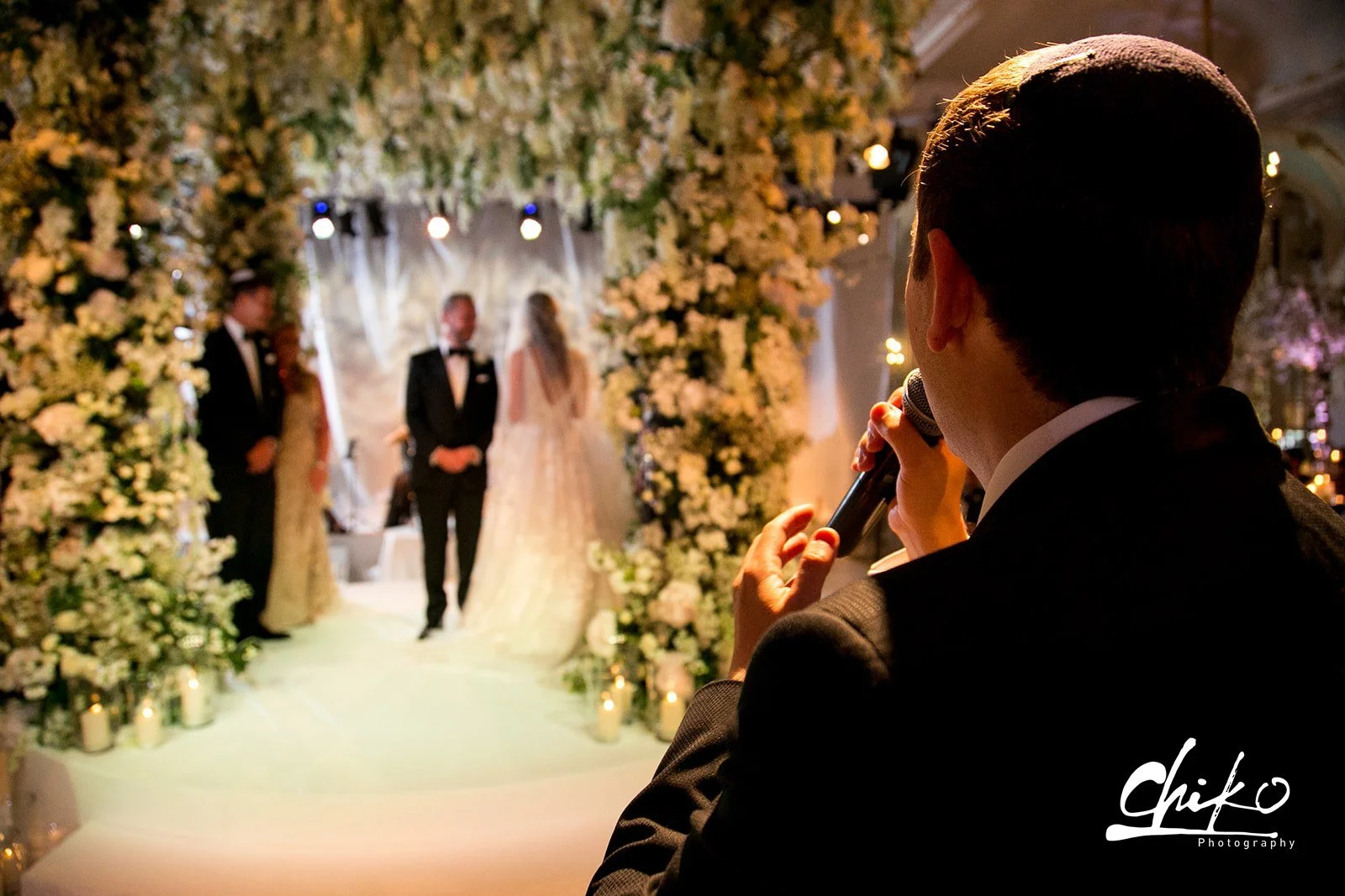 A man in a tuxedo is giving a speech with a microphone at a wedding ceremony. In the background, the bride and groom are standing under a floral arch. The setting is decorated with flowers and candles.