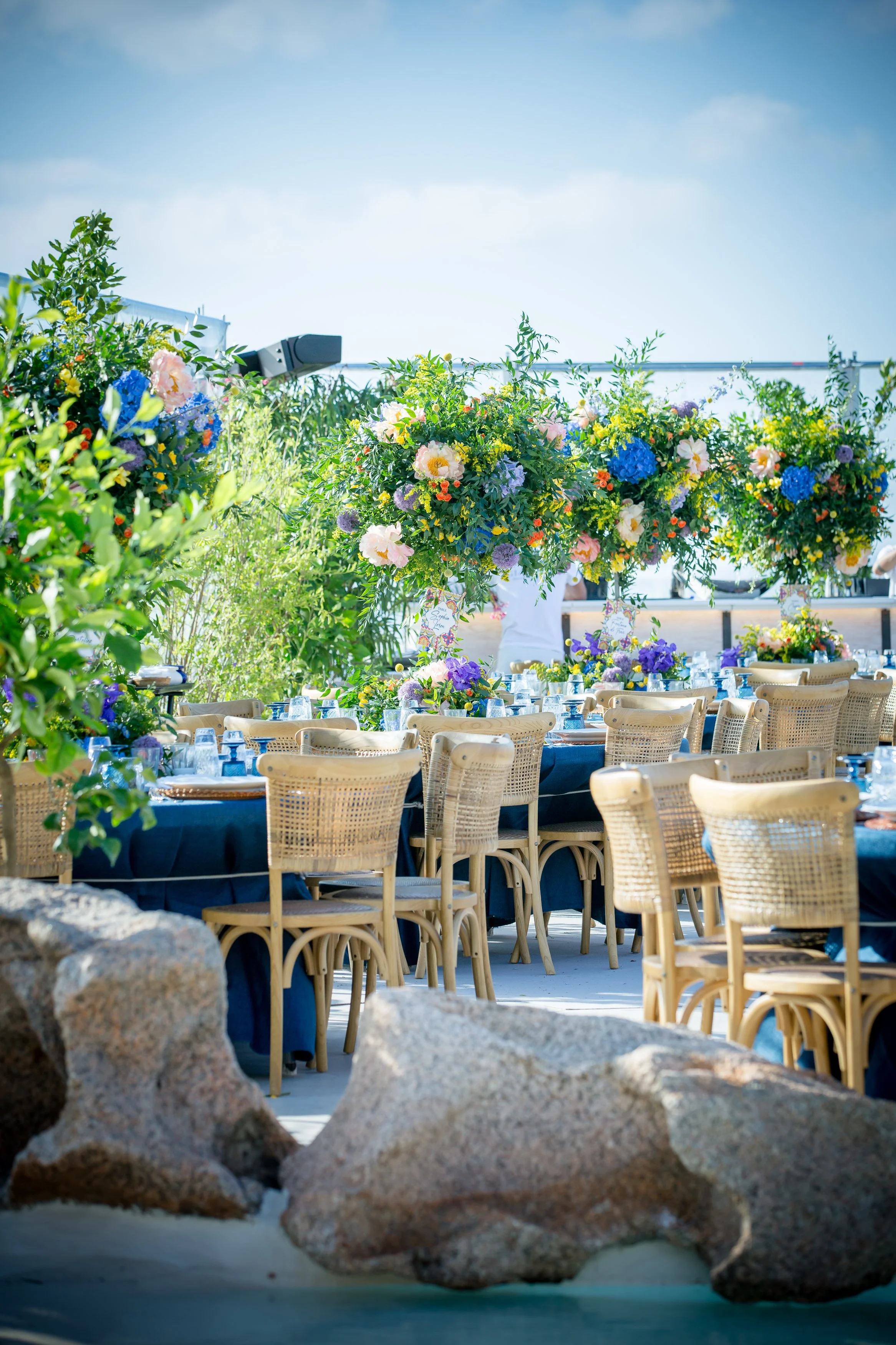 Outdoor event setup with tables and chairs decorated with flowers, surrounded by rocks, with a clear sky overhead.