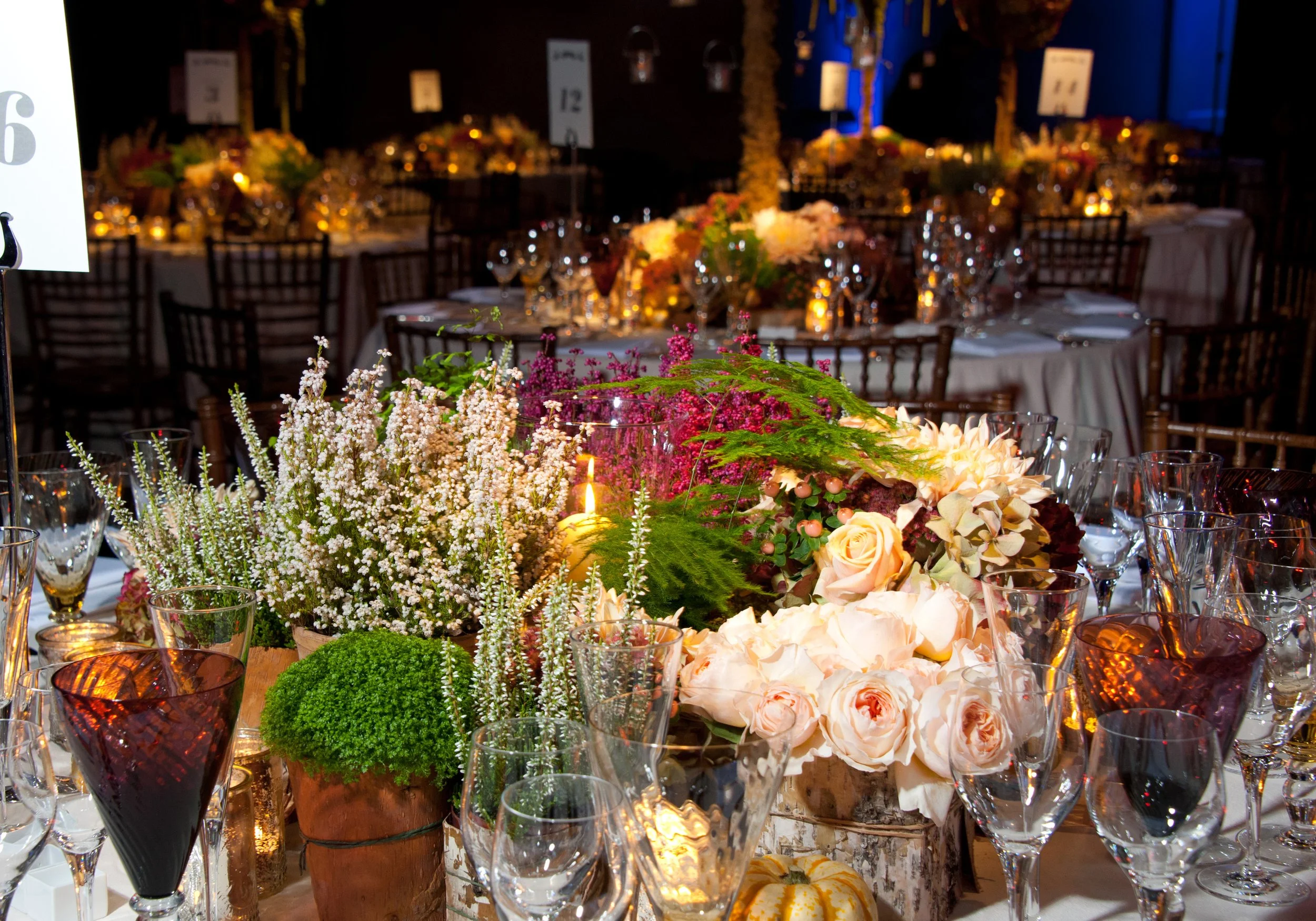 Elegant event table setup with floral centerpieces, surrounded by glassware and candles, in a dimly lit room with decorated tables in the background.