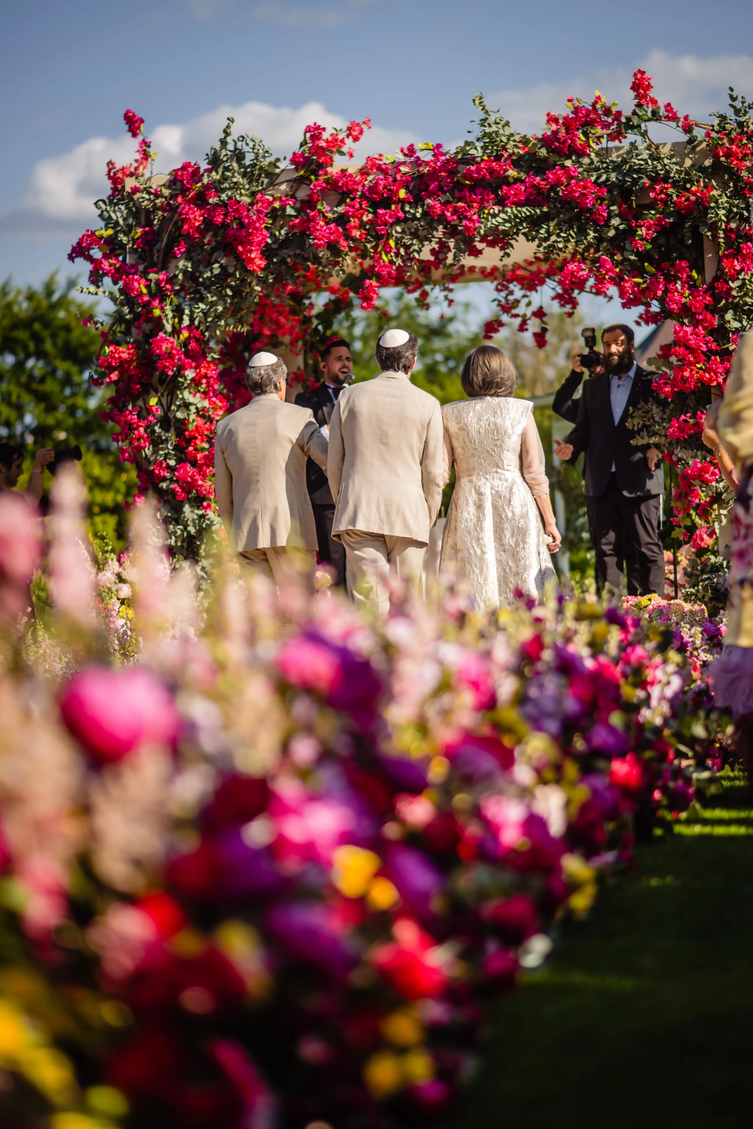 A wedding ceremony outdoors with a flower arch, couple and officiant, surrounded by guests and vibrant flowers. Fairmont Windsor Park