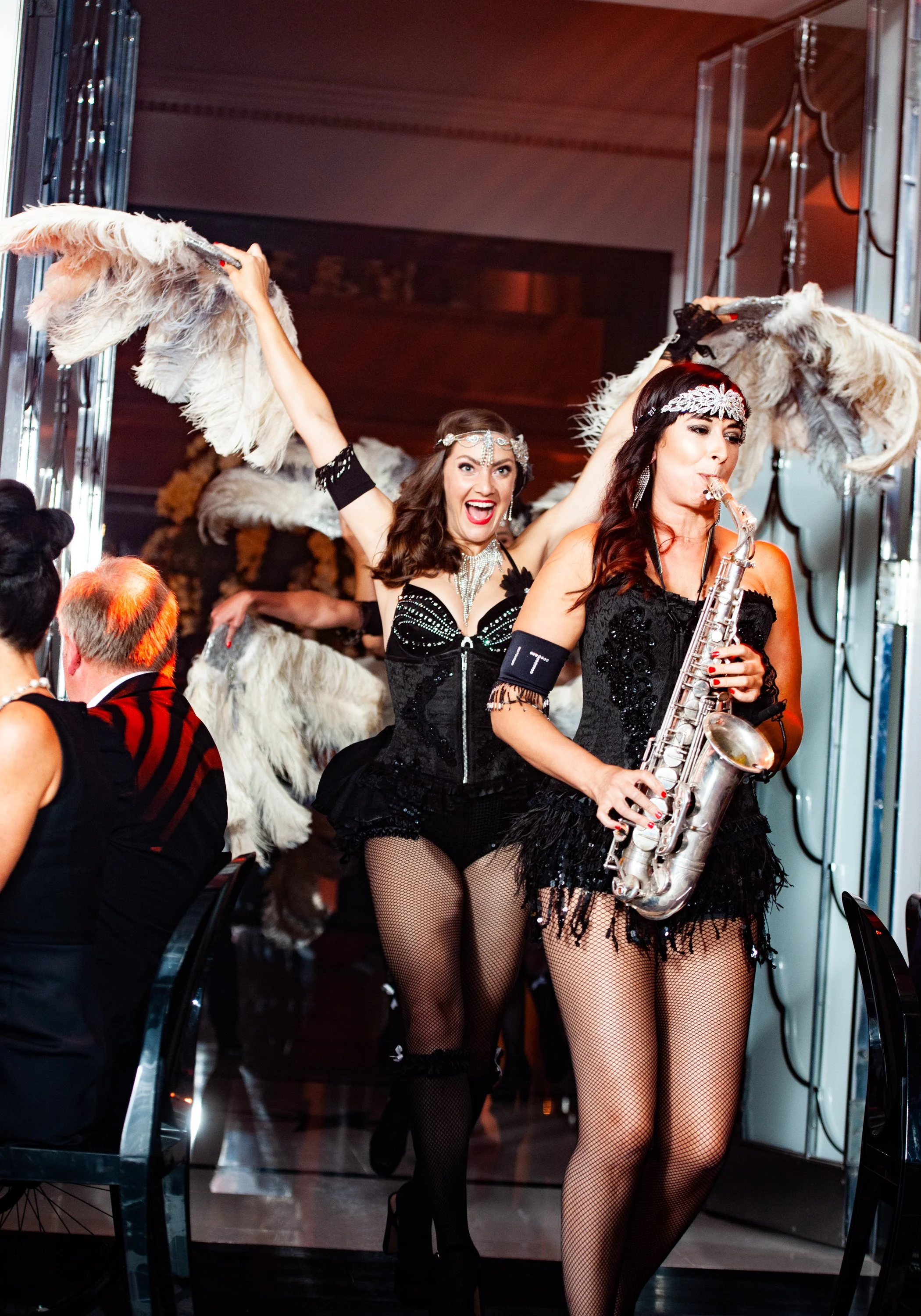 Two women in vintage showgirl costumes walking through a dimly lit room, one playing a saxophone and the other holding large feather fans, while guests seated at tables watch.