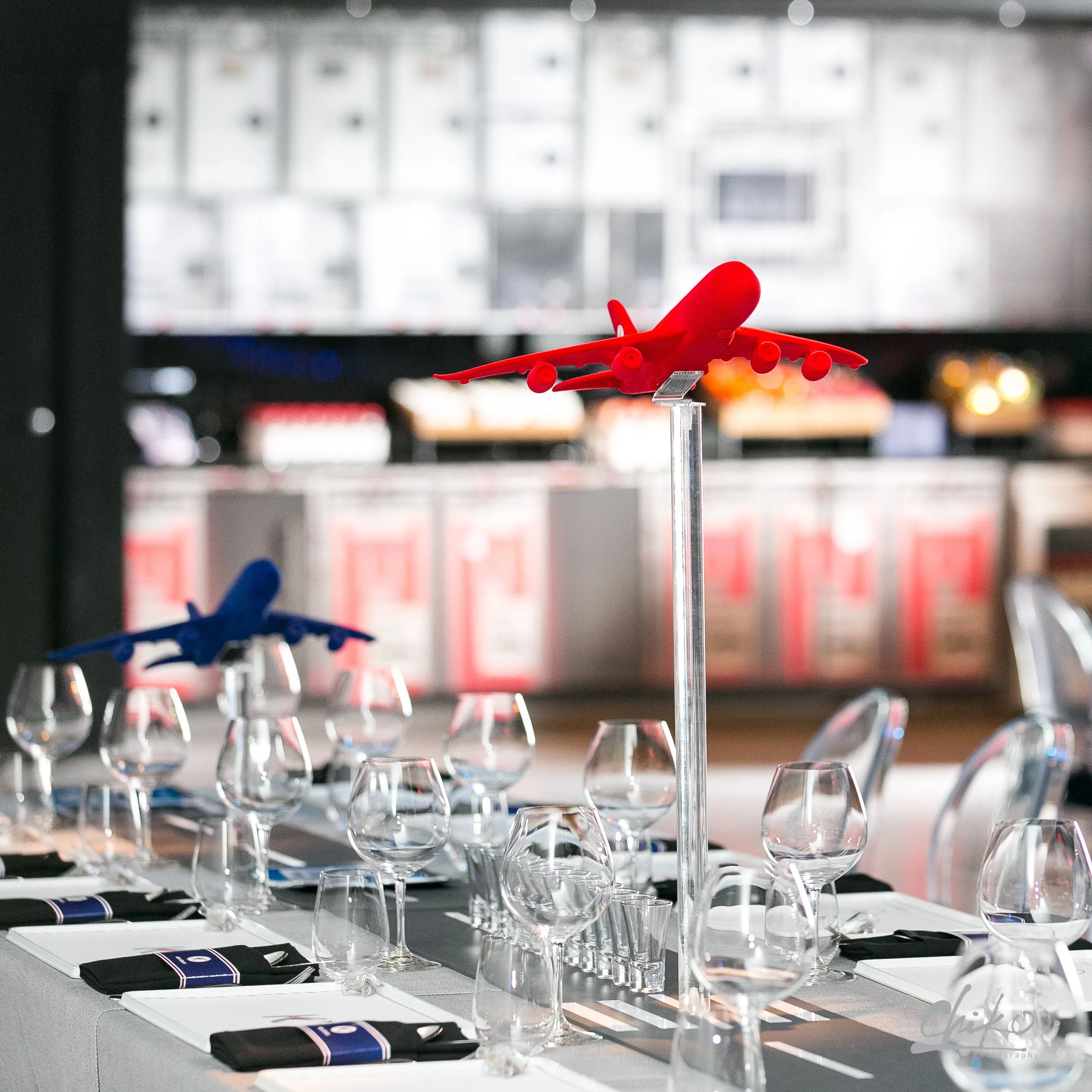 A miniature red airplane model on a table set with wine glasses, black napkins, and menus, in front of a blurred background of a larger screen or window showing buildings.