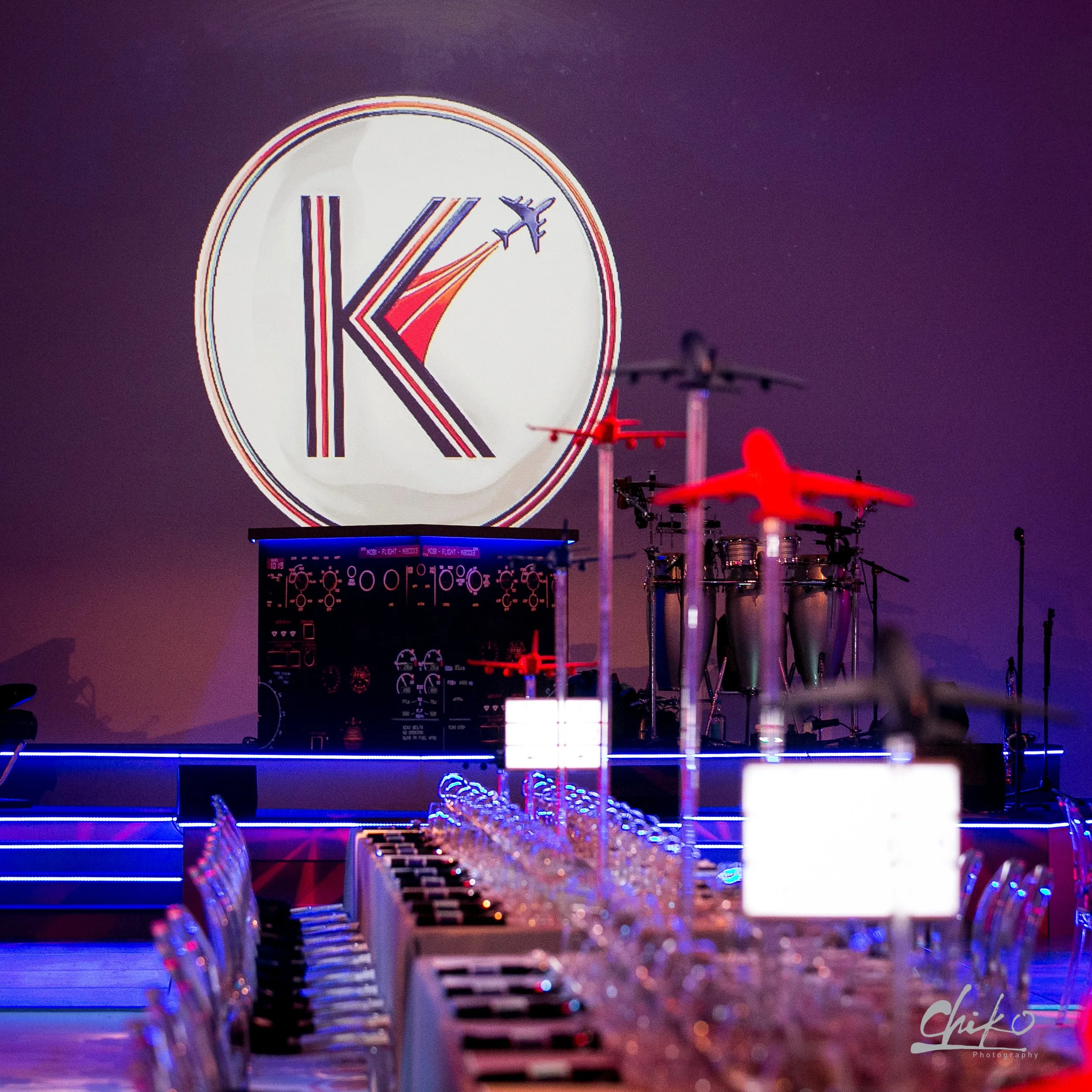 A large digital clock displays the time as 10:19, with an airplane icon next to the time. There are red and purple aircraft images in the background, and a table with glasses and a digital control panel in the foreground, all lit with purple and blue