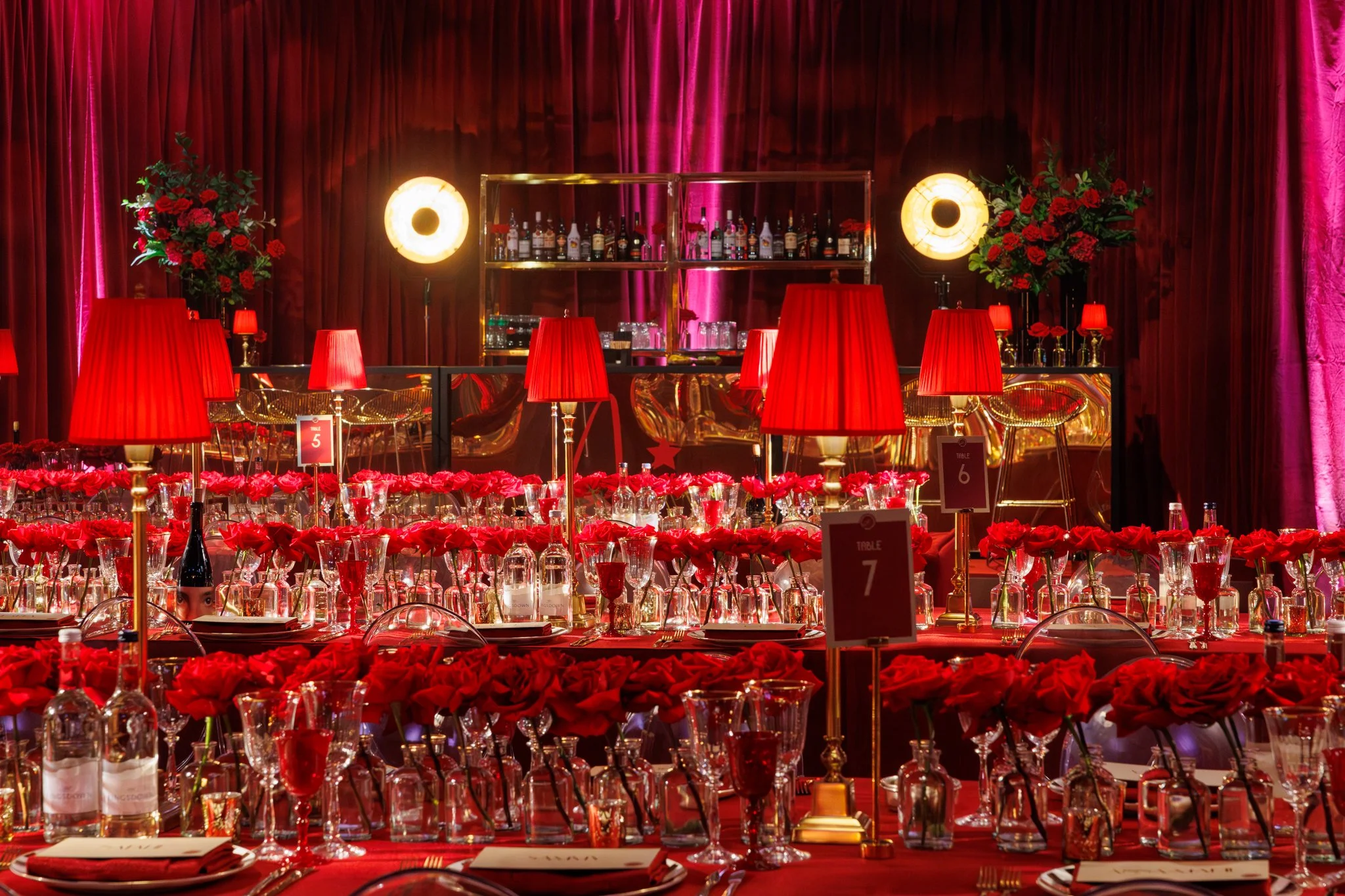 A decorated banquet hall with tables set for a formal event. The tables are adorned with red flowers, glassware, and table number cards. The background features warm lighting, red curtains, and a bar area with bottles and barware.