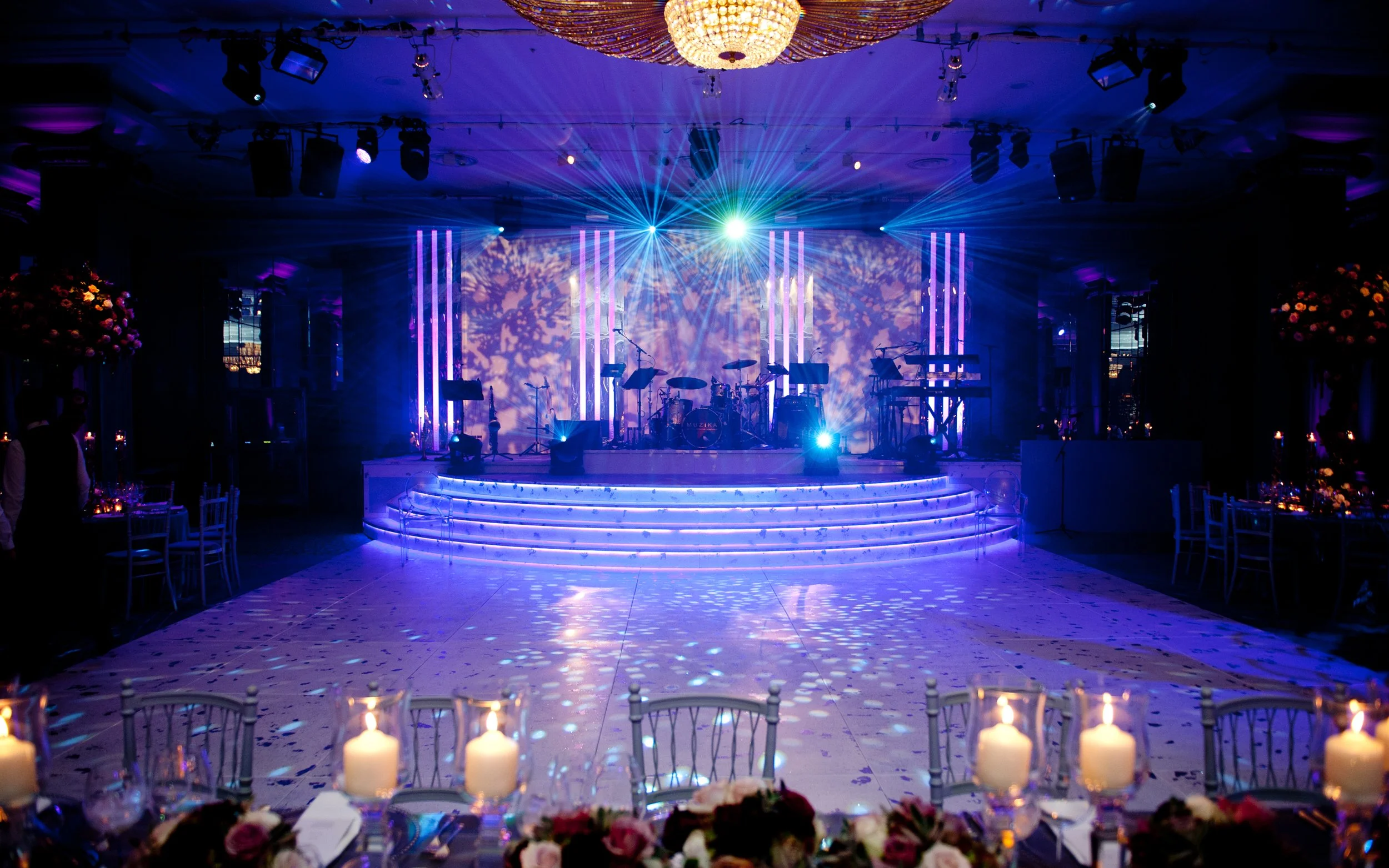 A decorated event stage with colorful lighting effects, musical instruments, and a chandelier overhead, set for a wedding at Grosvenor House