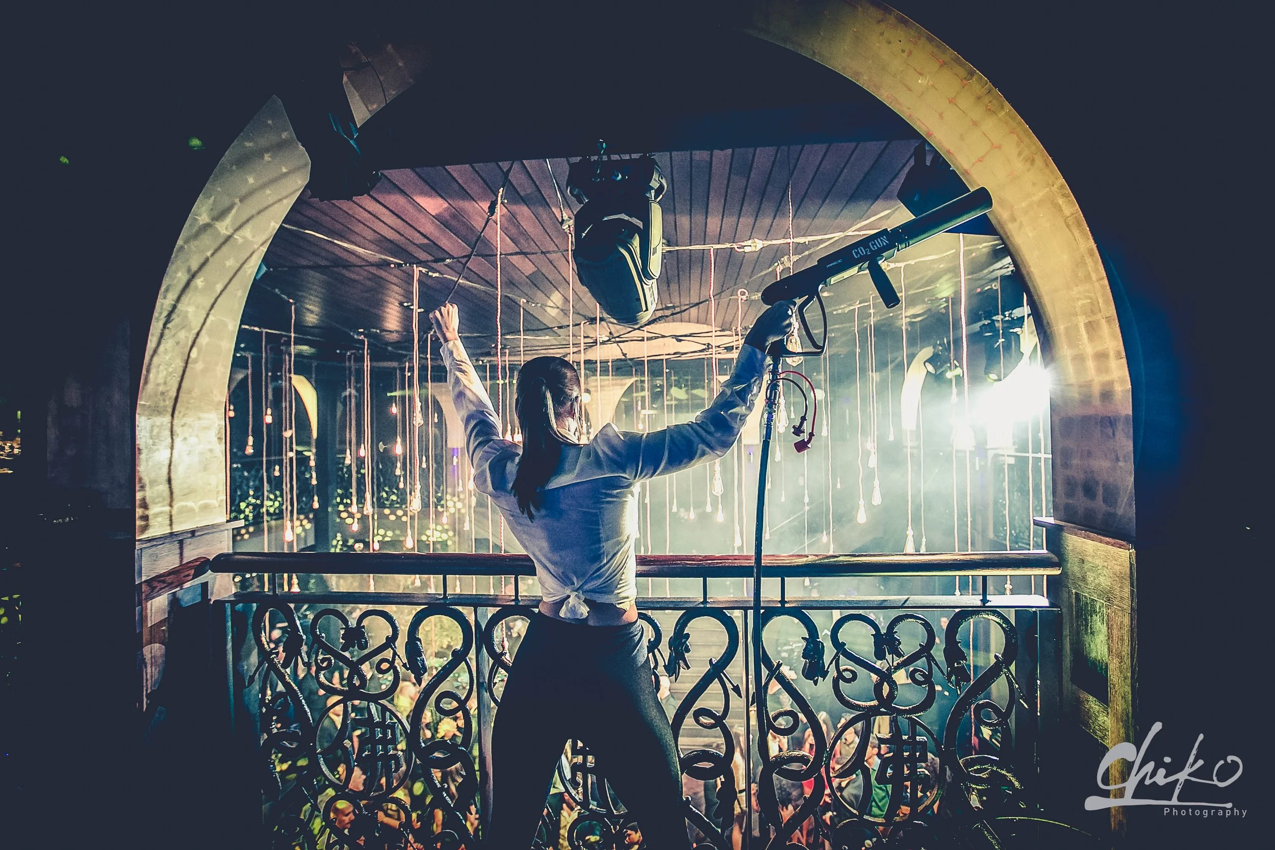 A woman with tied back hair standing on a balcony with ornate railing at night, holding up her right hand, while a professional camera is directed toward her, recording or photographing her. The background shows an interior with hanging light bulbs a