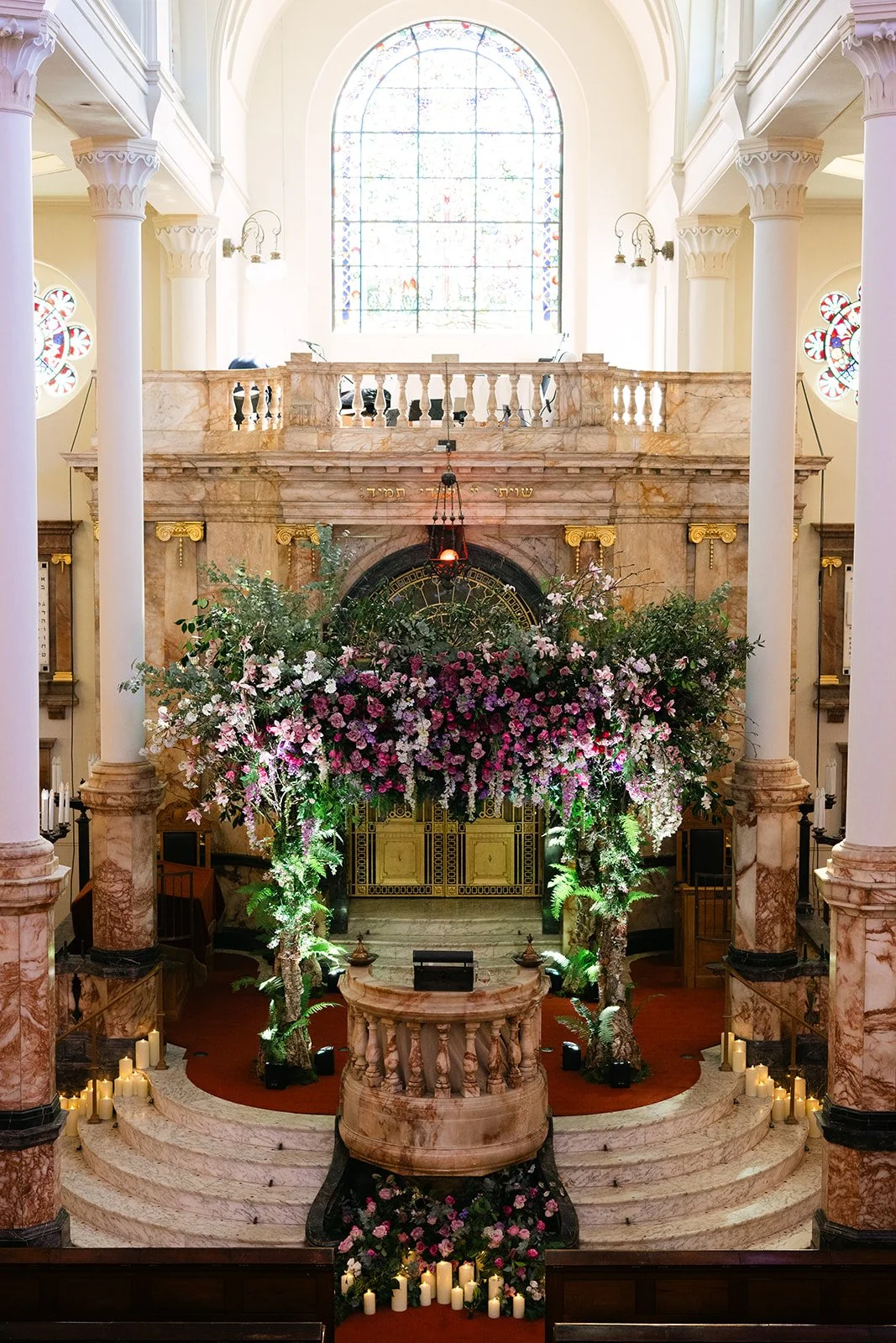 Interior of a church or cathedral decorated with a large floral arch and numerous candles, with stained glass windows and marble columns. Wedding at The Peninsula London