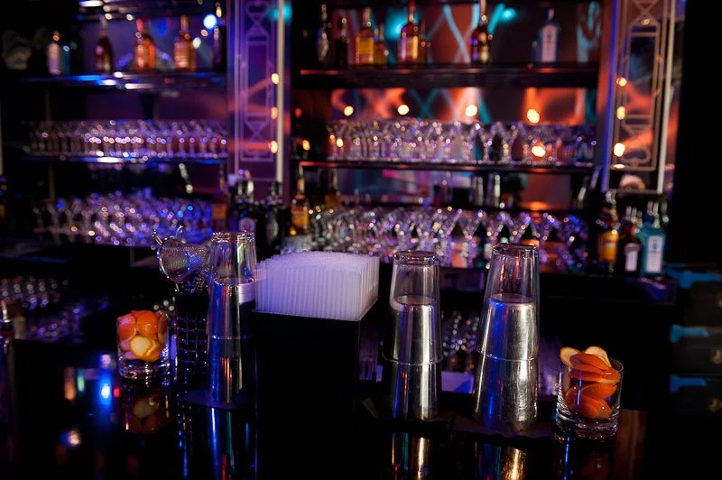 Bar counter with glasses, cocktail napkins, and assorted liquor bottles displayed in the background.