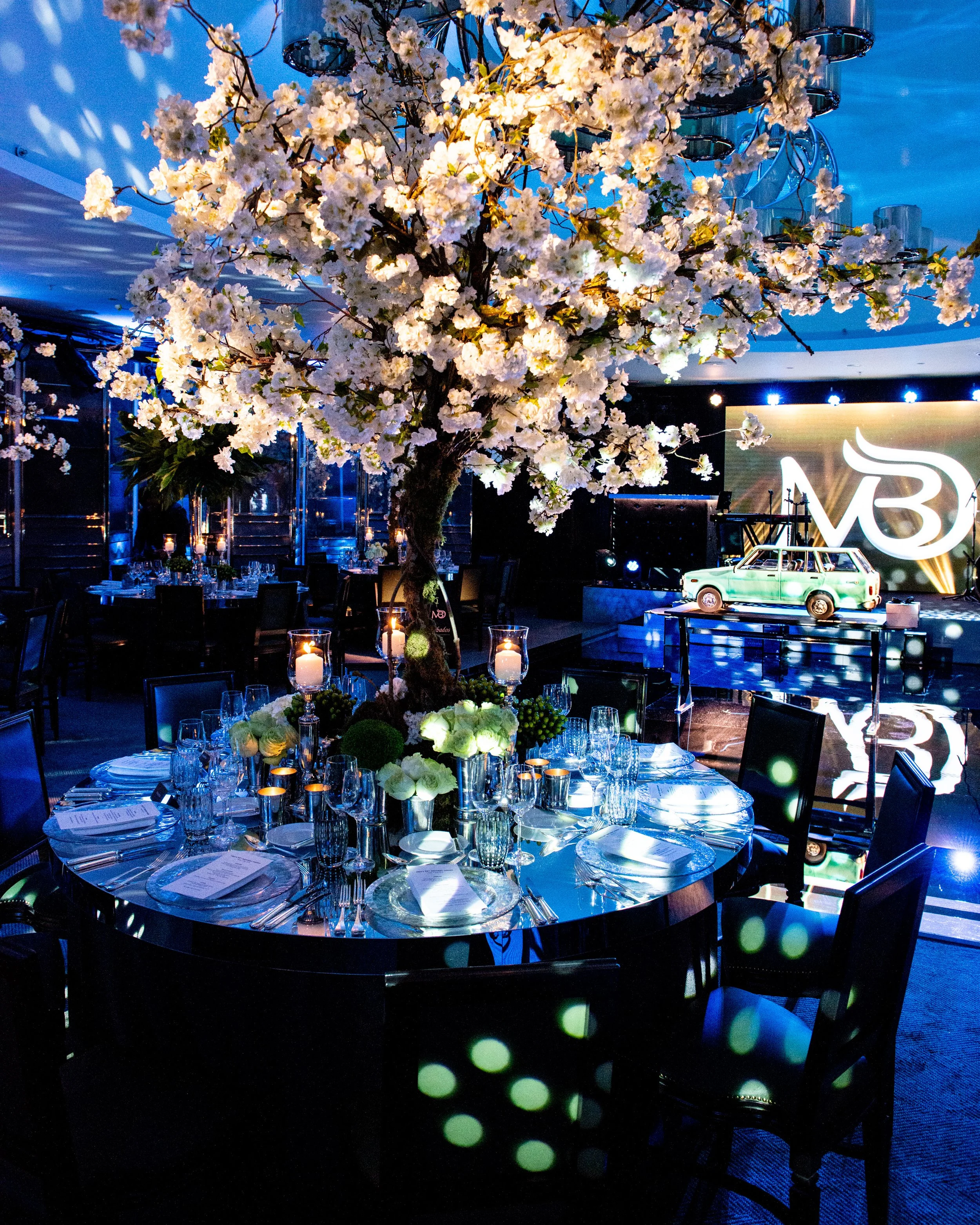 A round banquet table decorated with flowers, candles, and place settings, illuminated by blue lighting, in an event hall with a large floral centerpiece of white flowers, and a stage featuring the logo 'V3' and a decorative car prop.