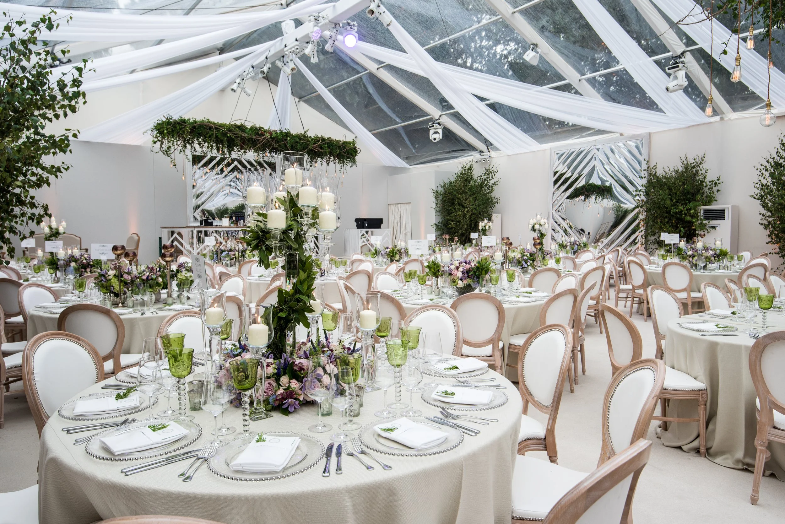 Elegant event setup inside a transparent tent with draped white fabric ceiling, round tables with white tablecloths, floral centerpieces, and green glassware, surrounded by comfortable chairs, and decorated with candles and greenery.