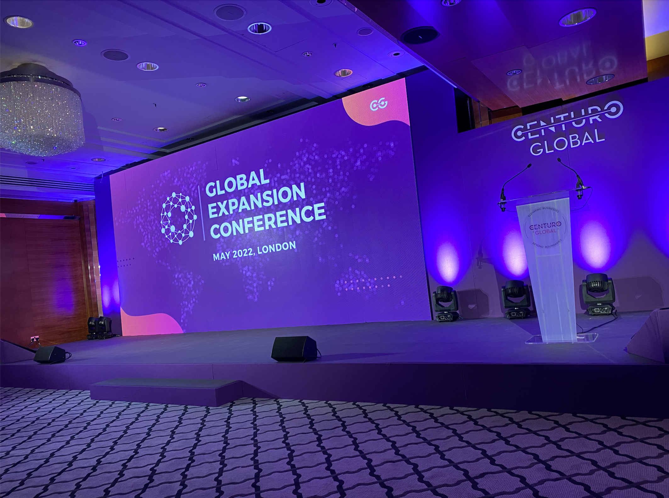 Stage setup at the Global Expansion Conference in London, May 2022, with a large screen displaying the event name and a podium with a banner, ambient blue and purple lighting, and a chandelier in a conference hall.