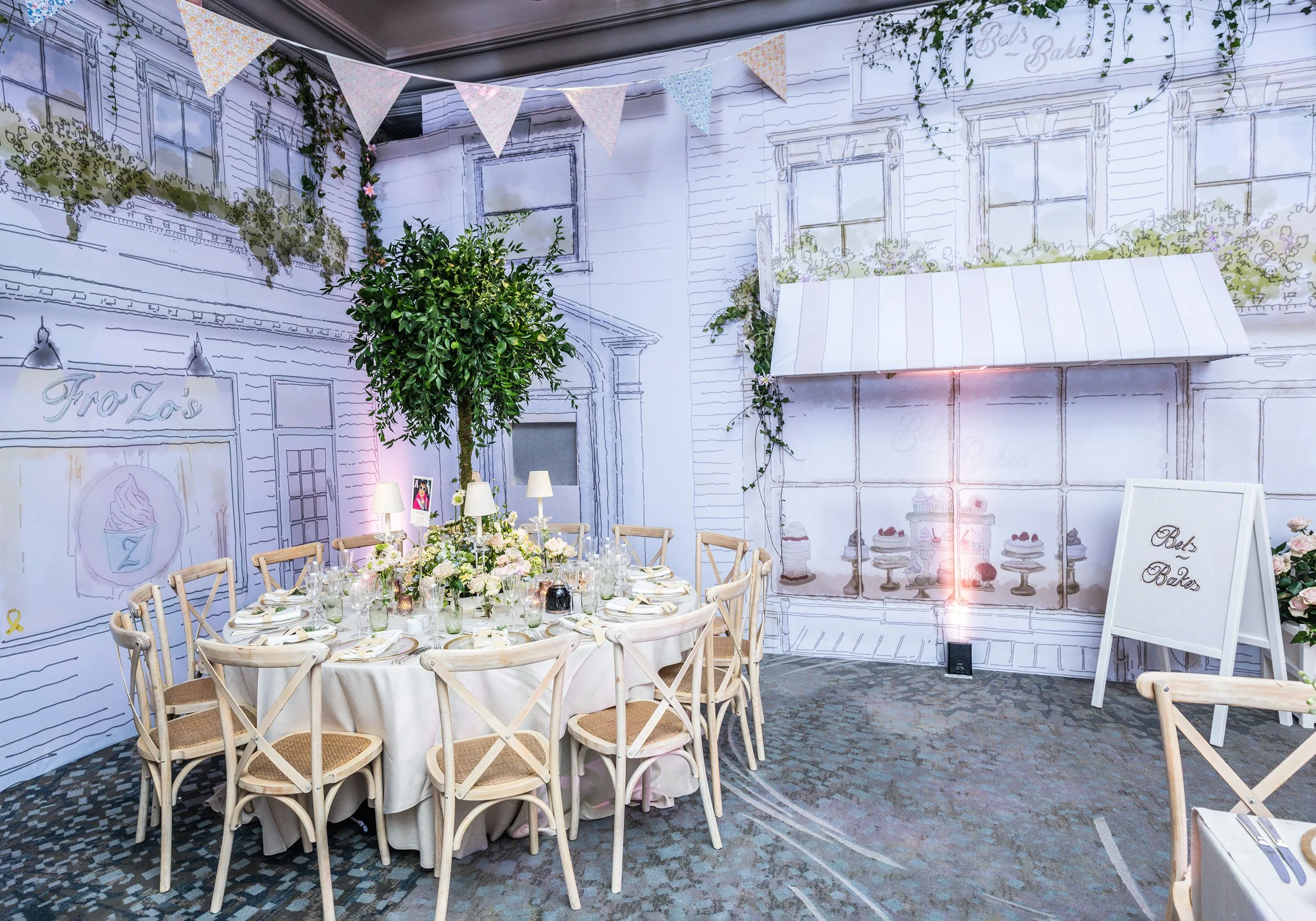 Decorative indoor scene with a large circular table set for a meal, surrounded by wooden chairs. The background features a wall with line drawings of buildings, windows, and floral elements, and is decorated with hanging bunting flags and greenery