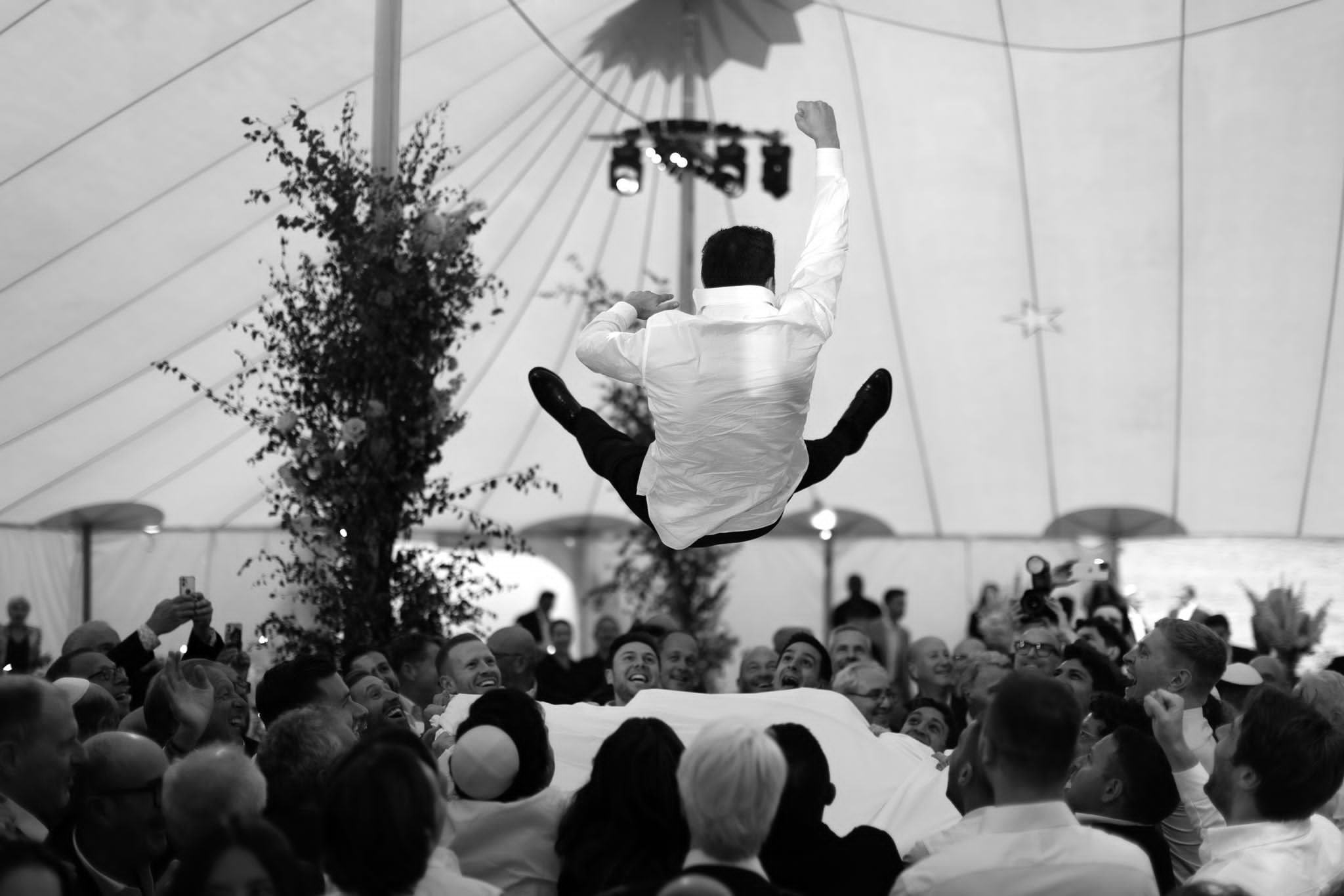 A man in a white shirt is performing a backflip in front of a large crowd inside a tent, with some audience members taking pictures. The scene is in black and white.