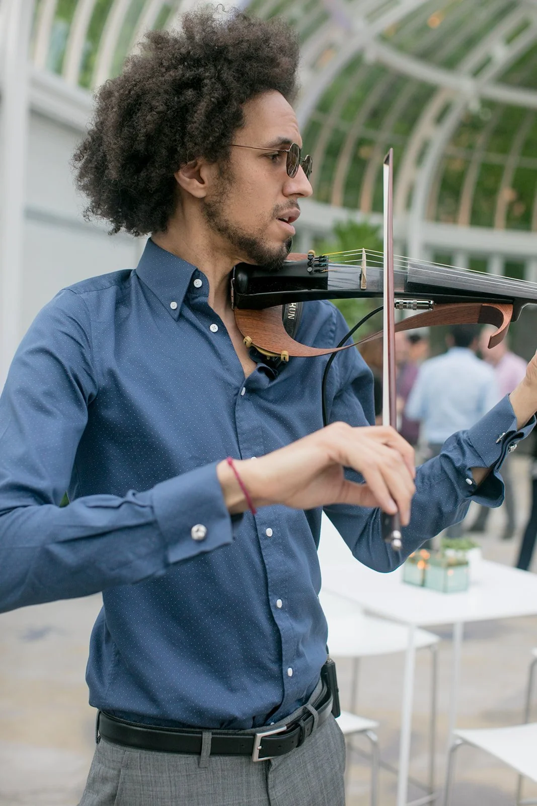 Man playing a violin with a bow in an outdoor setting, wearing sunglasses, a blue shirt, and gray pants.