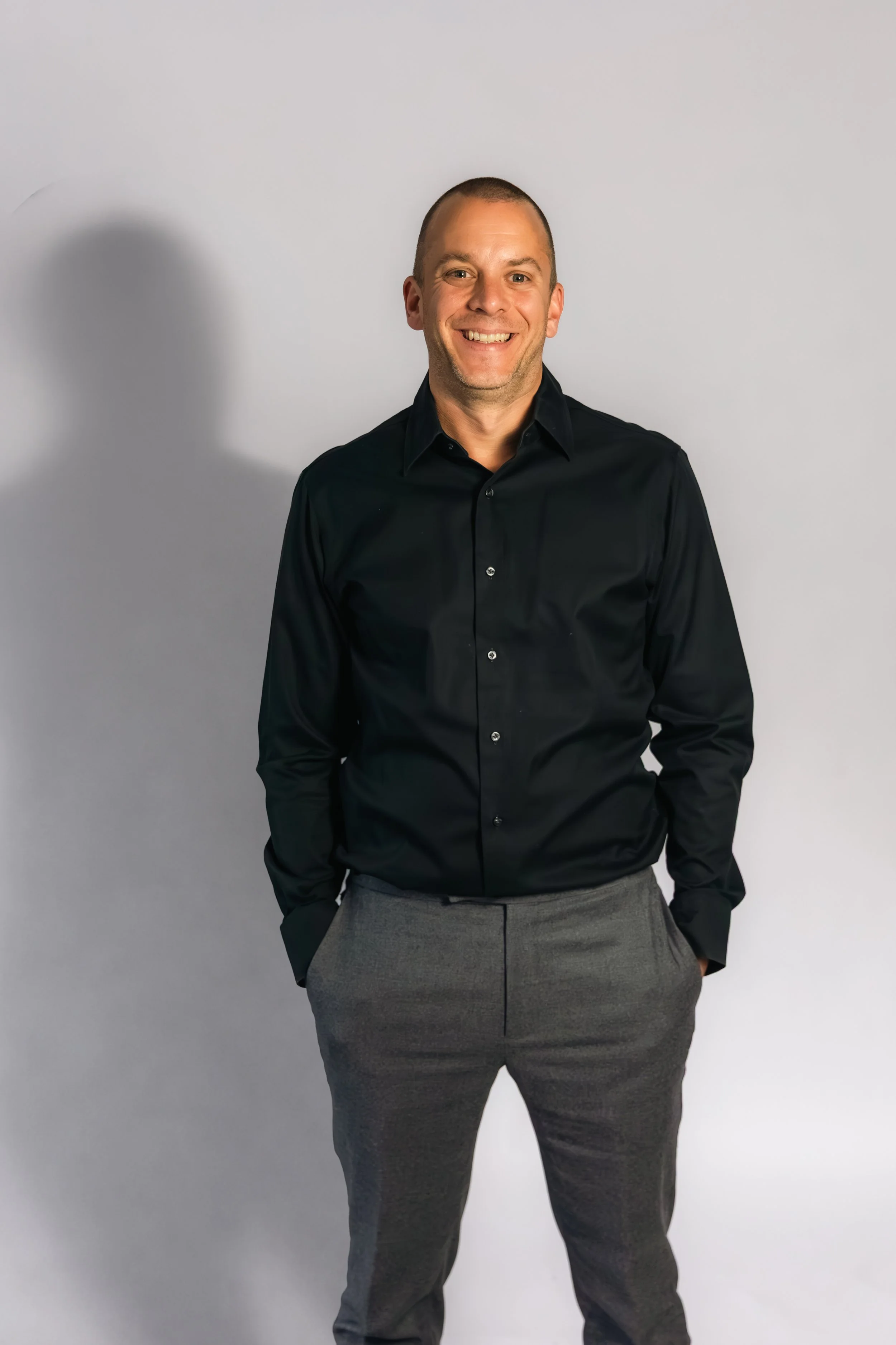 Laurie Weitzkorn smiling man stands against a plain light gray background, wearing a dark button-up shirt and gray trousers, with his hands in his pockets.