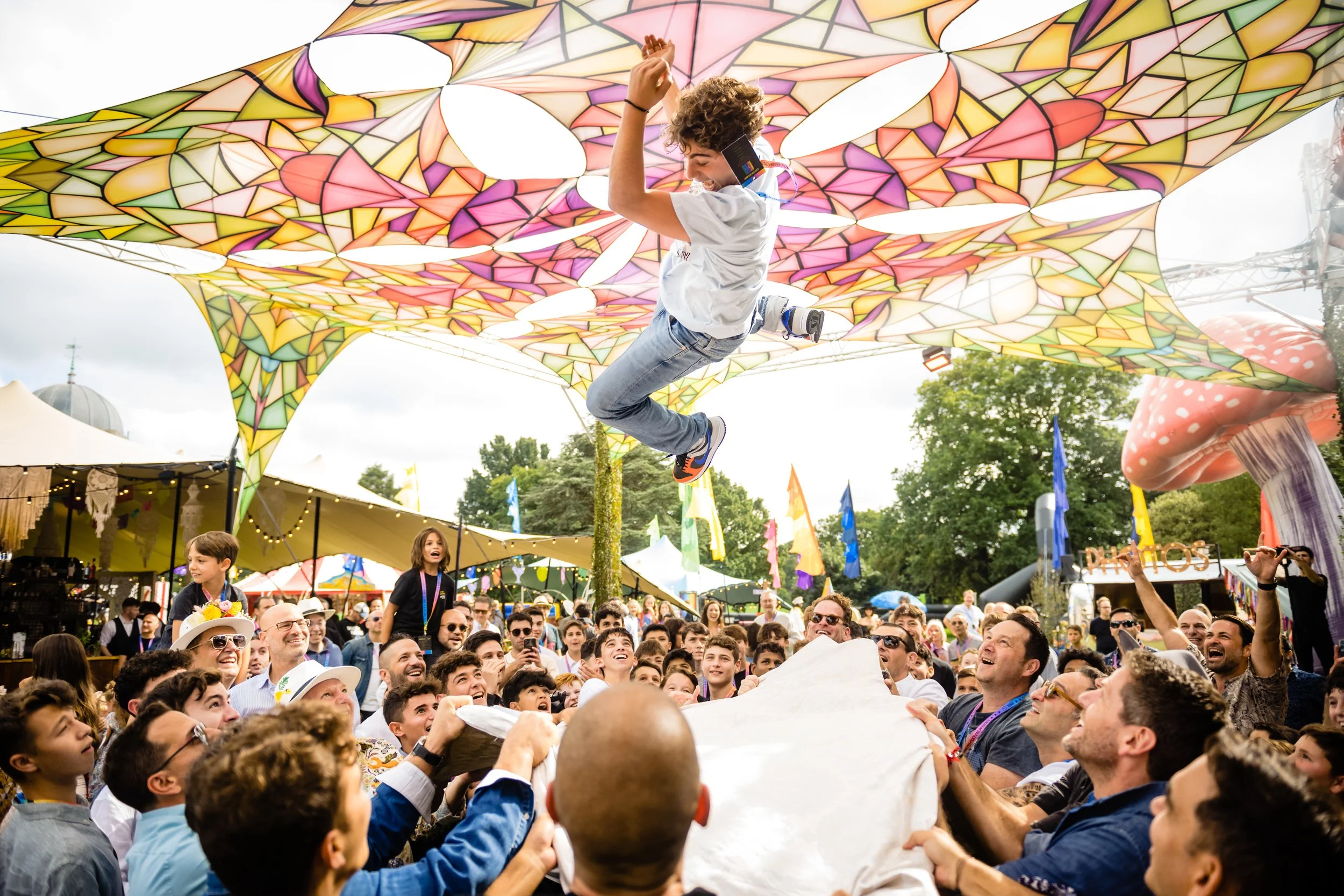A person is high above a crowd at an outdoor festival, jumping on a large, colorful parachute-like fabric canopy with stained-glass pattern. The crowd looks up and smiles, enjoying the event.