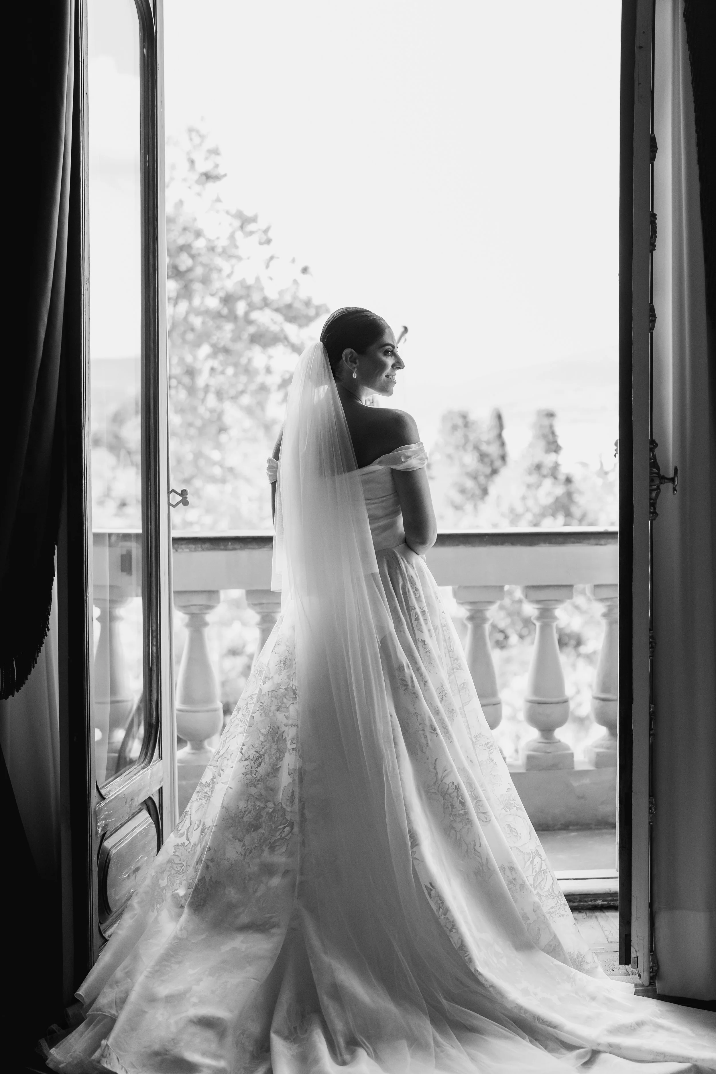A bride standing by an open door leading to a balcony, wearing a wedding dress and veil, looking sideways with a smile, with trees visible outside in black and white at Villa Palmieri