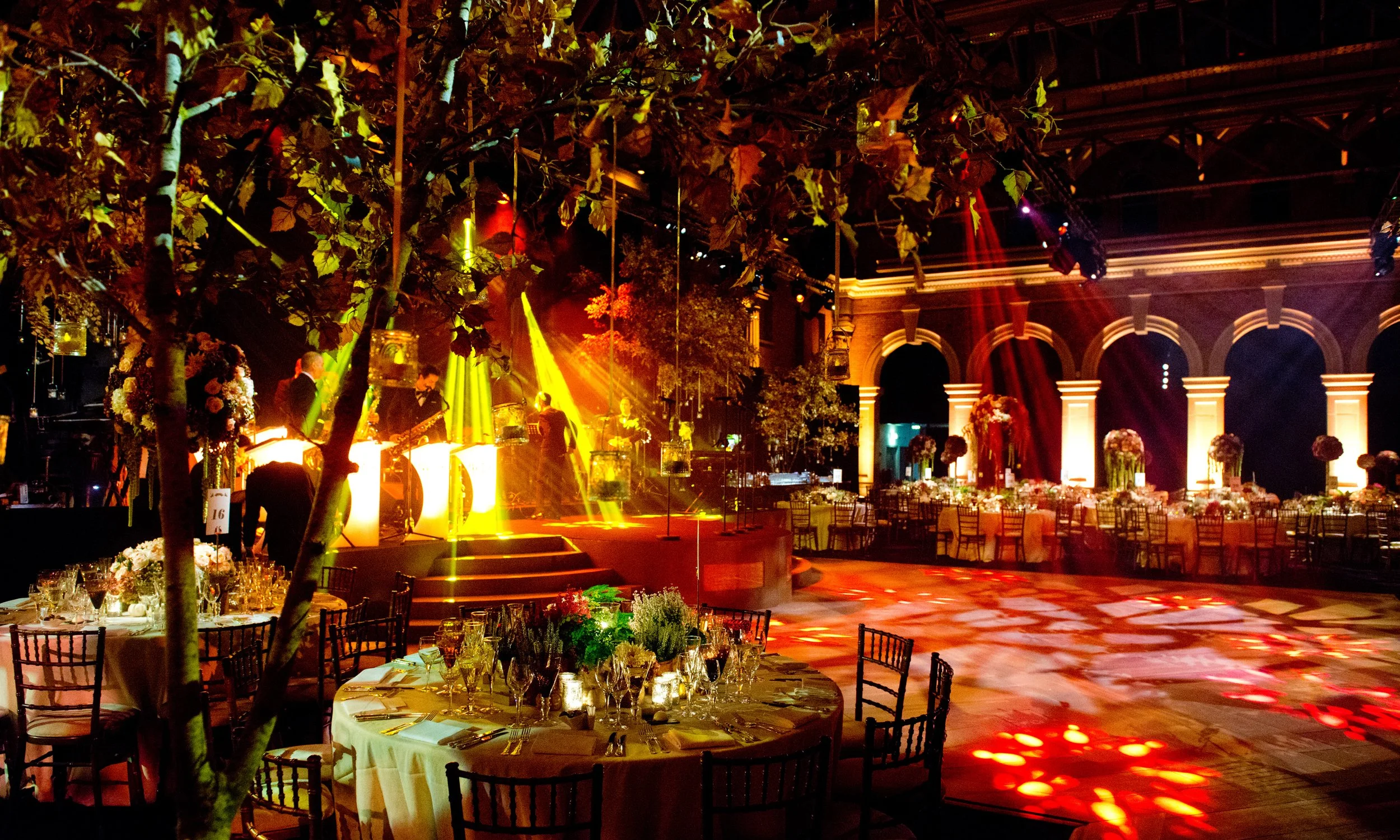 Elegant banquet hall decorated for a formal event with tables set with flowers and glassware, and a live band playing on a stage with colorful lighting.
