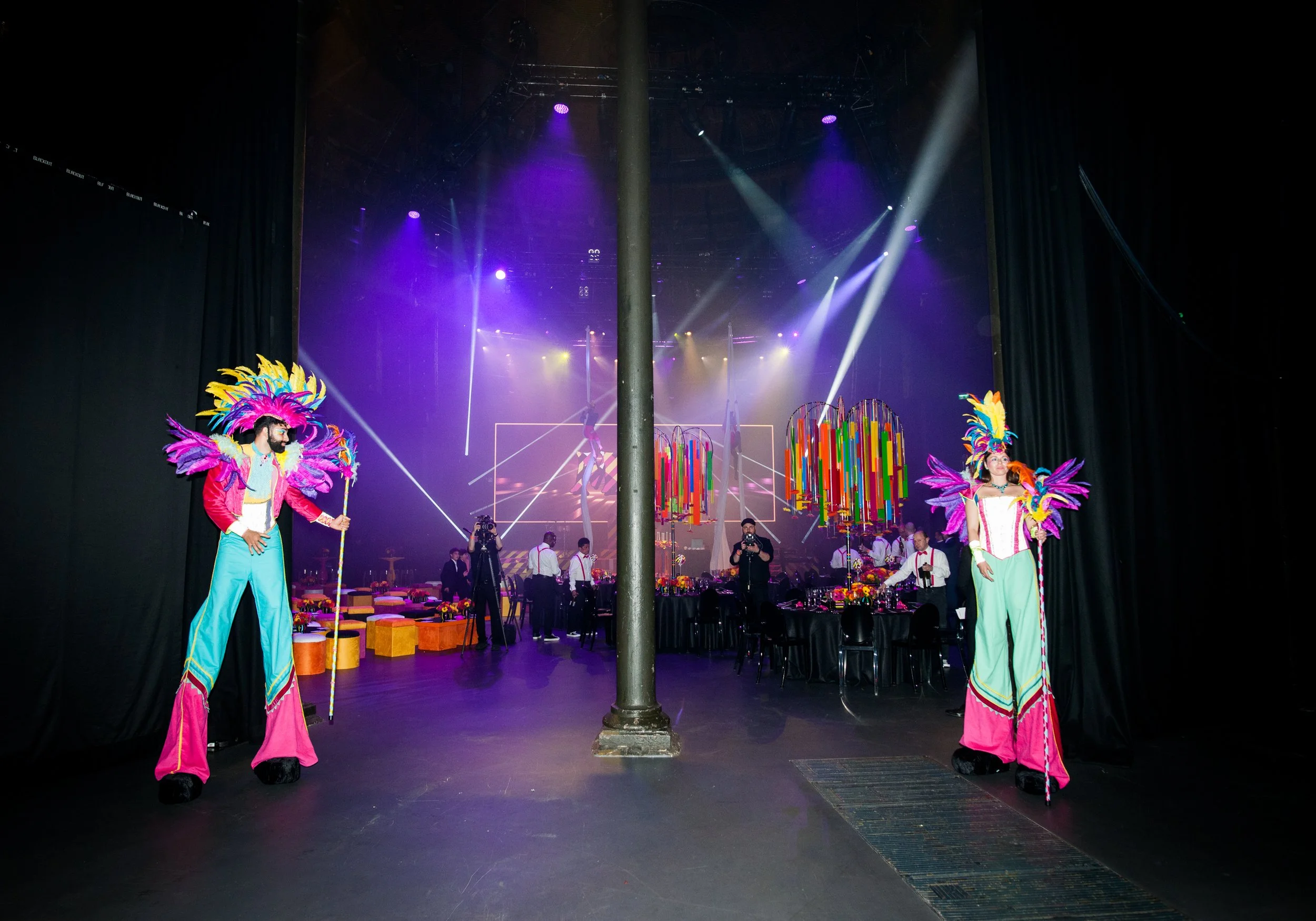 Two performers in colorful costumes with feathered headdresses and long capes standing on stage, with a decorated venue and stage lights in the background at the roundhouse