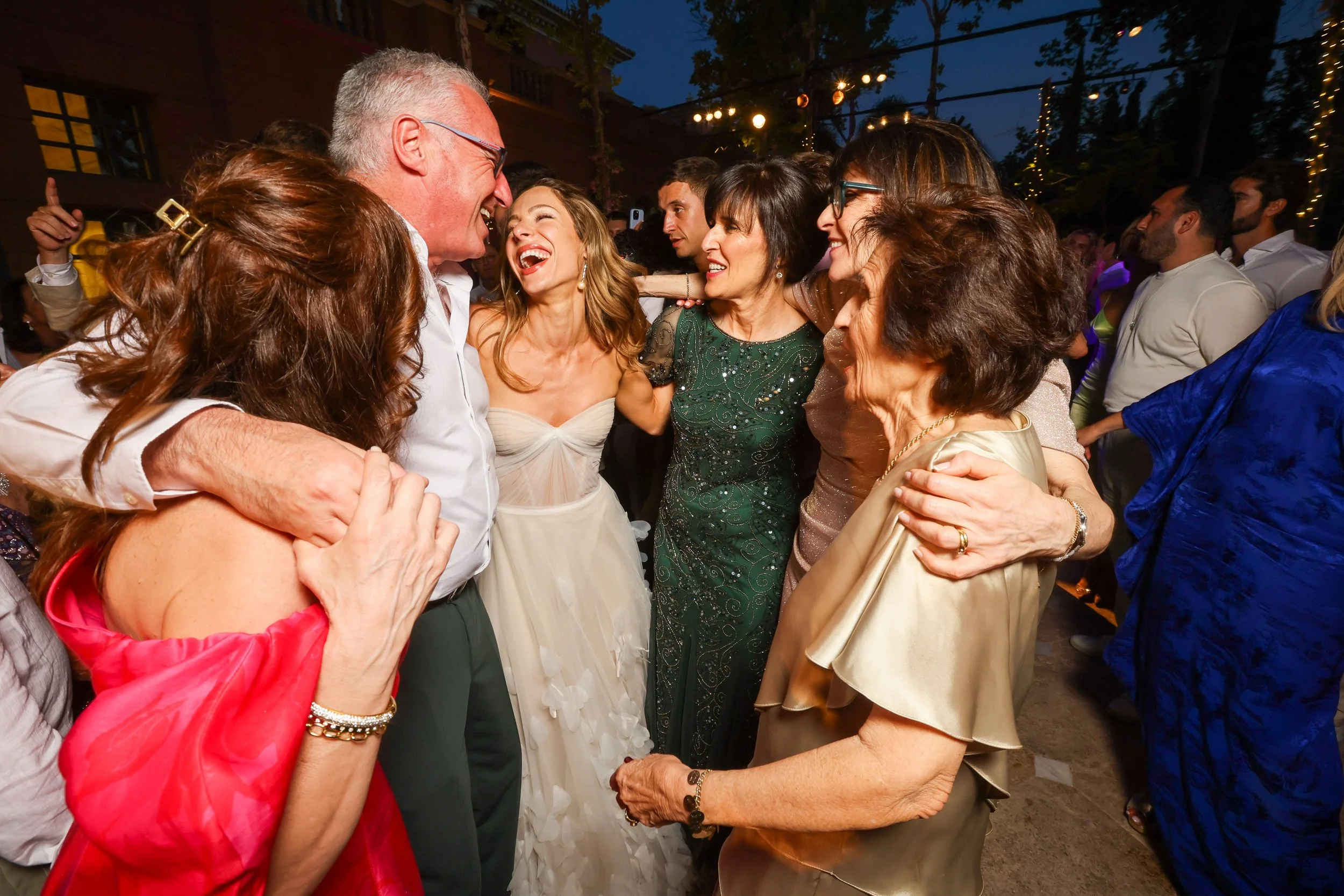 People dancing and celebrating at a party, smiling and enjoying each other's company in an outdoor setting with string lights and trees.