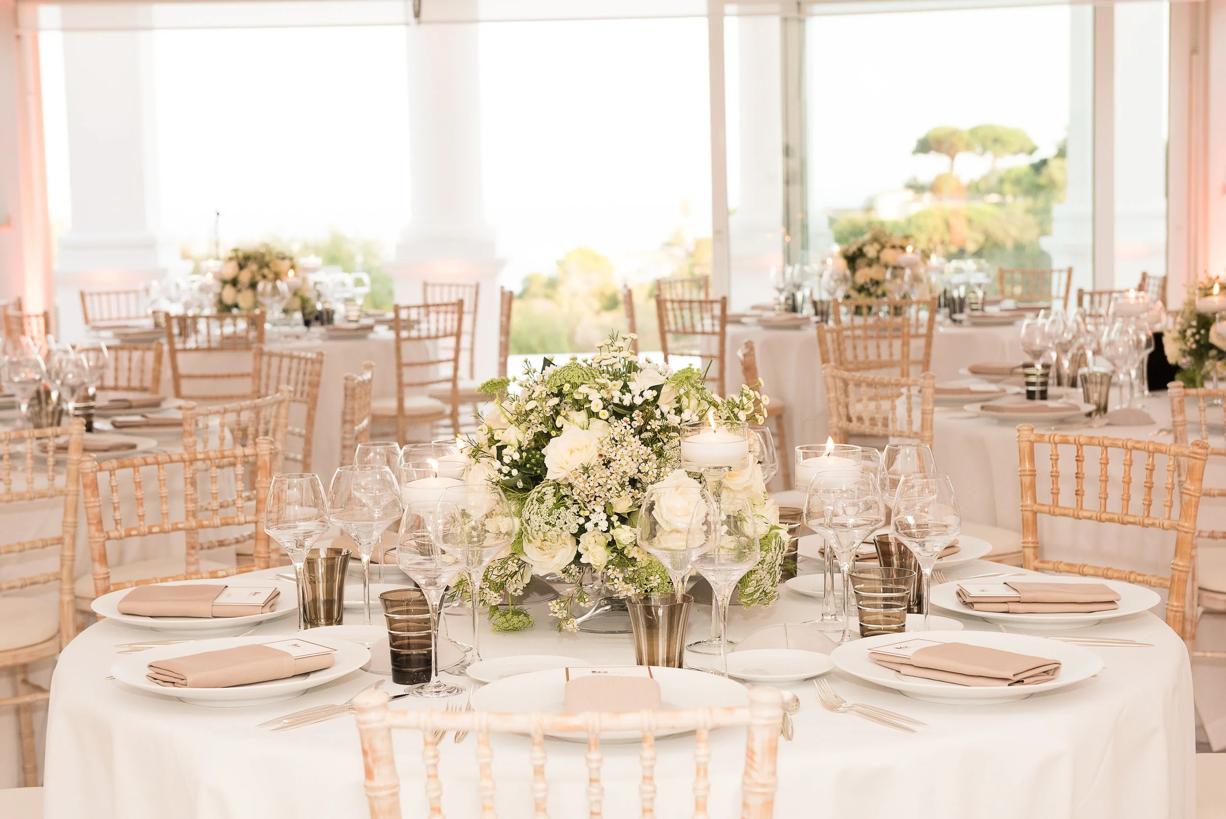 Elegant reception table set with beige napkins, glassware, and a floral centerpiece featuring white roses and greenery in a bright, airy room with large windows and wooden chairs.