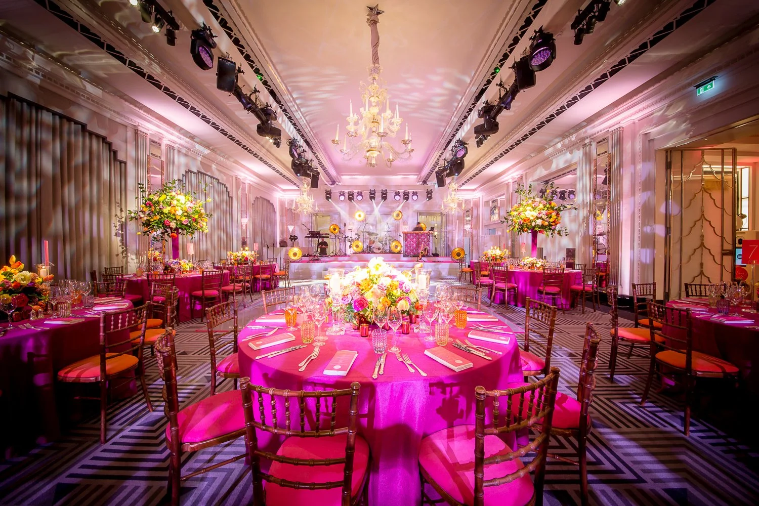 Elegant banquet hall decorated with pink tablecloths, floral centerpieces, and chandeliers, prepared for a formal event with a stage and musical instruments at the front.