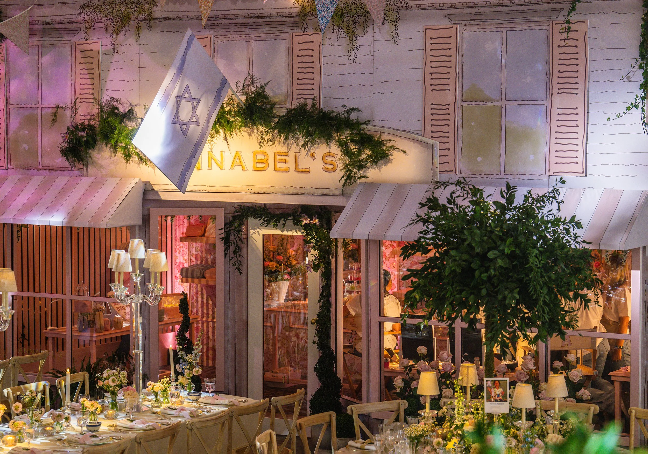 A Bat Mitzvah at Grosvenor House with the theme of Hamptons with floral arrangements, tables set with glassware and candles, adorned with greenery and with pink and yellow drapery, bunting, and string lights.