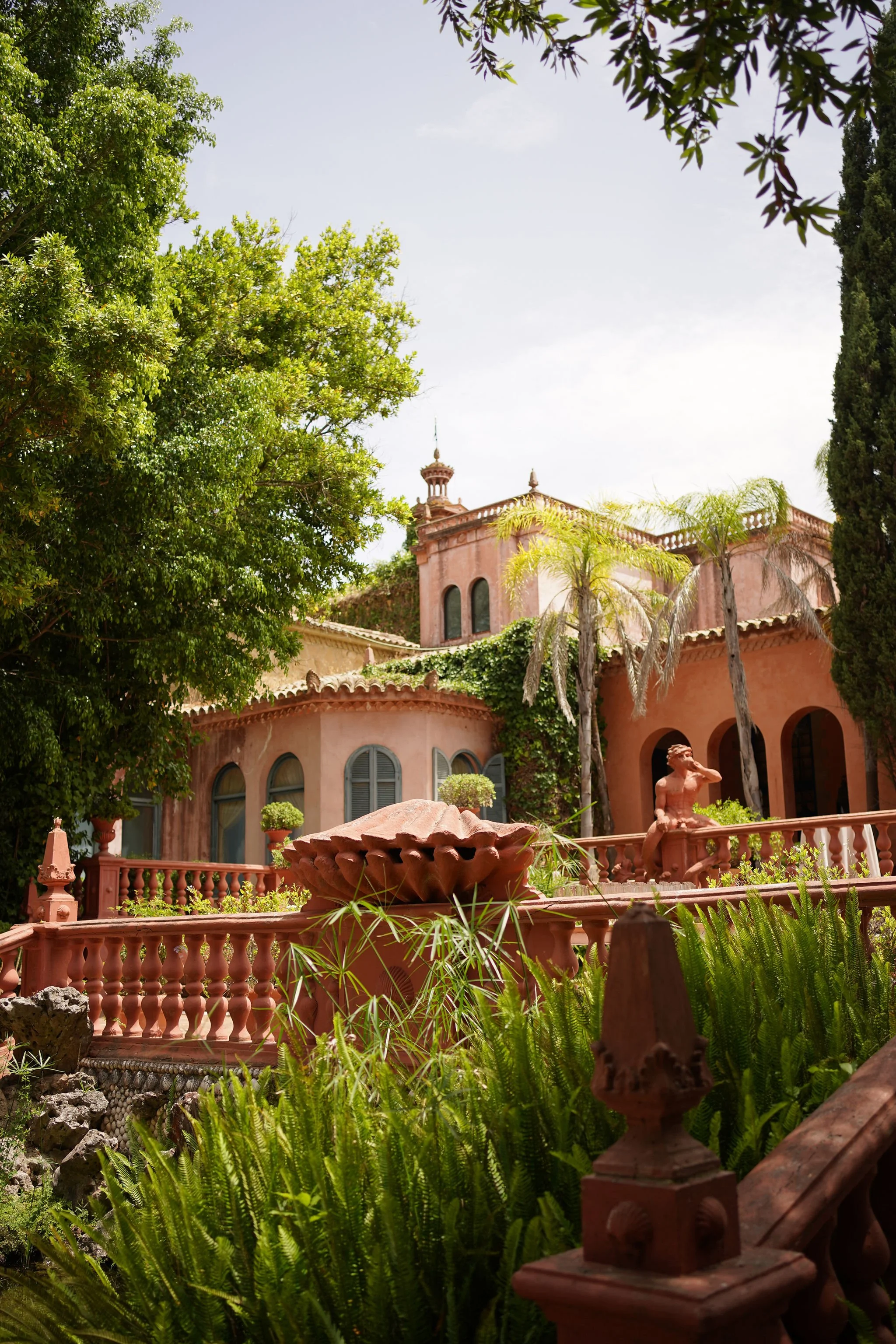 A garden scene with lush green trees and plants, a pinkish building with arched windows, a small tower, and a decorative fountain or structure in the foreground at Anantara Villa Padierna Resort in Marbella