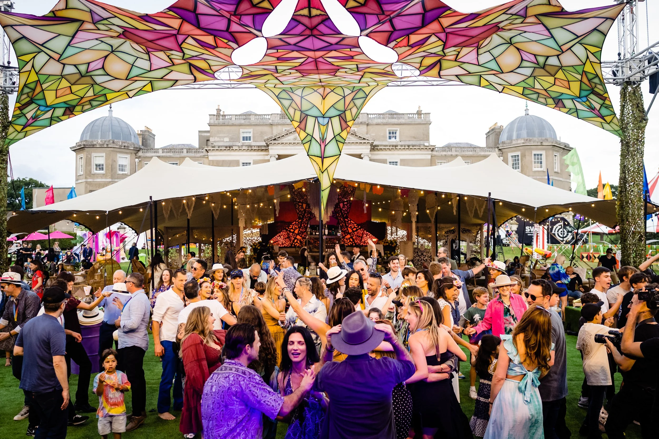 Crowd of people at an outdoor event with a large, colorful stained glass tent overhead and historic building in the background at a Bar Mitzvah at  Wrotham Park