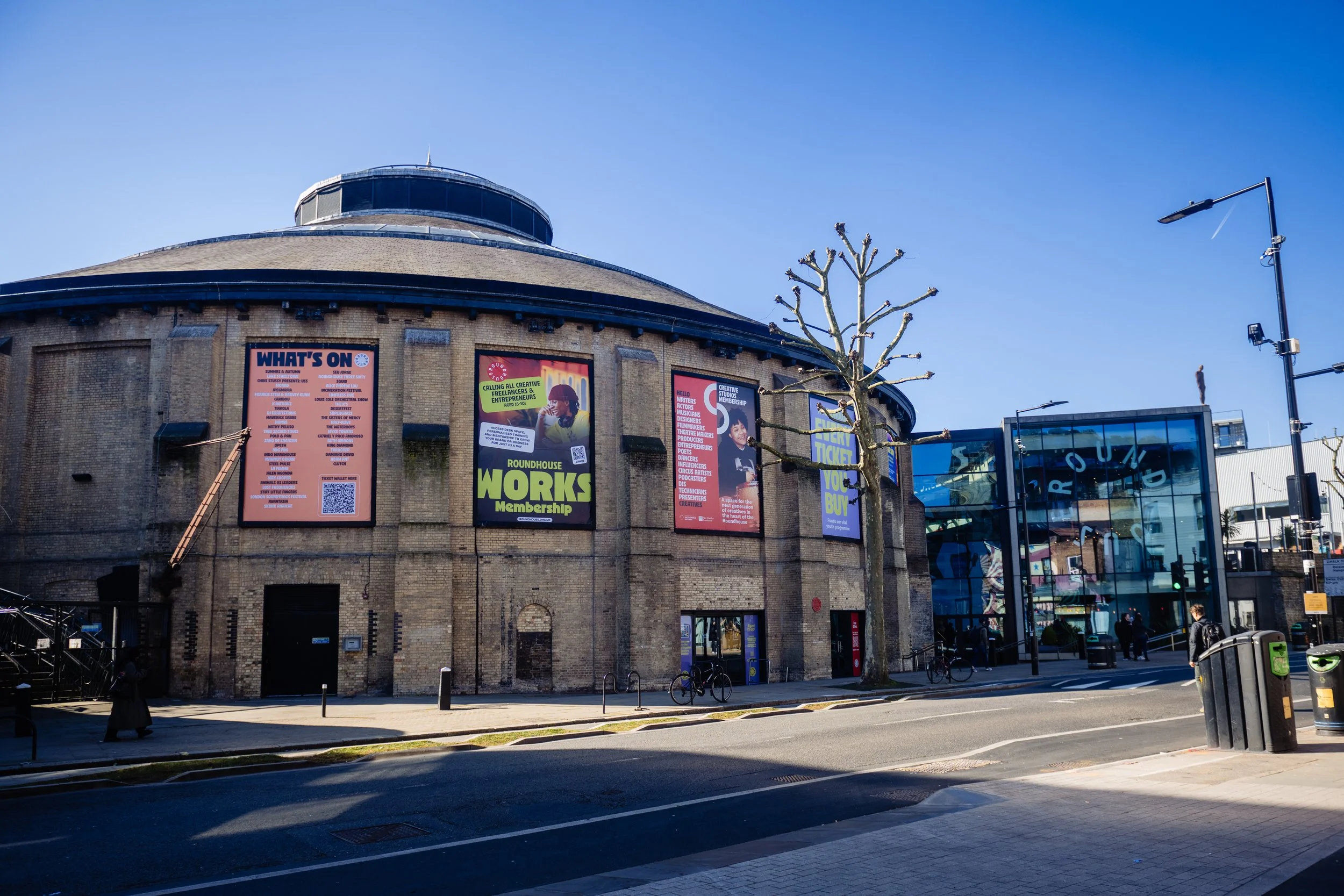 The Roundhouse and its brick building with event posters on the exterior wall, adjacent to a modern glass building, bicycle racks, and a few people walking on the sidewalk under a clear blue sky.