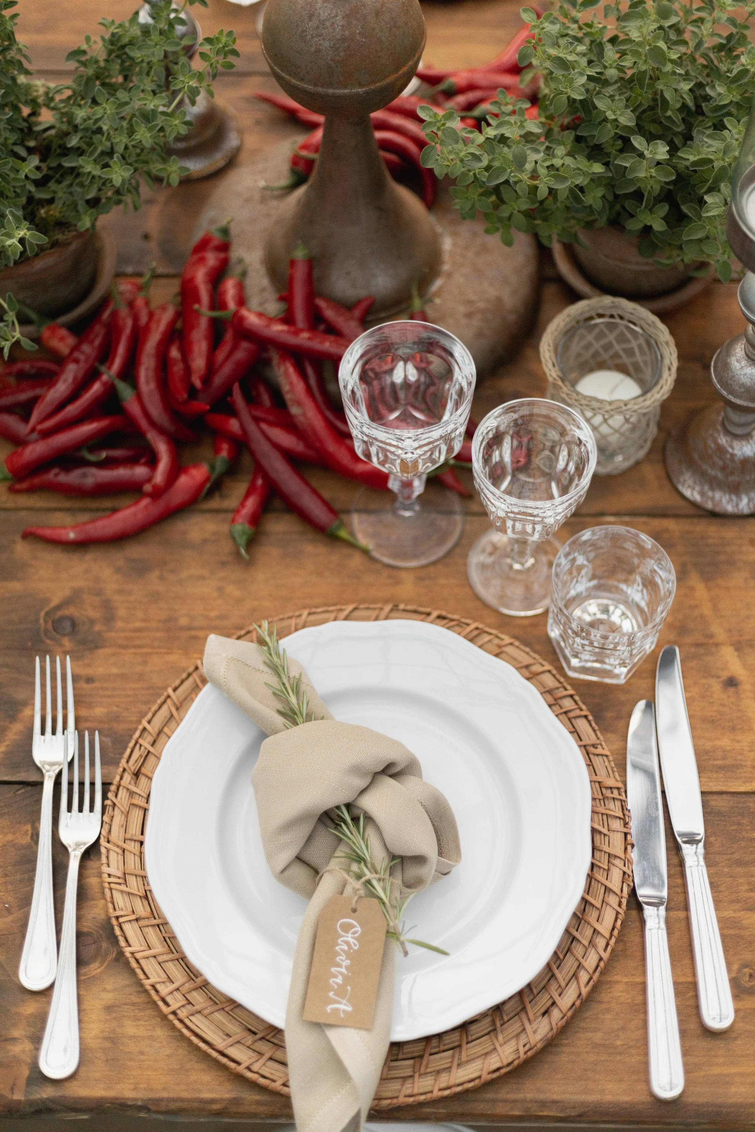 A rustic wooden table decorated for a meal with a white dinner plate on a woven placemat, a beige cloth napkin with a sprig of rosemary, and clear glassware. There are also potted green plants, red chili peppers, candles, and metal candlesticks in th