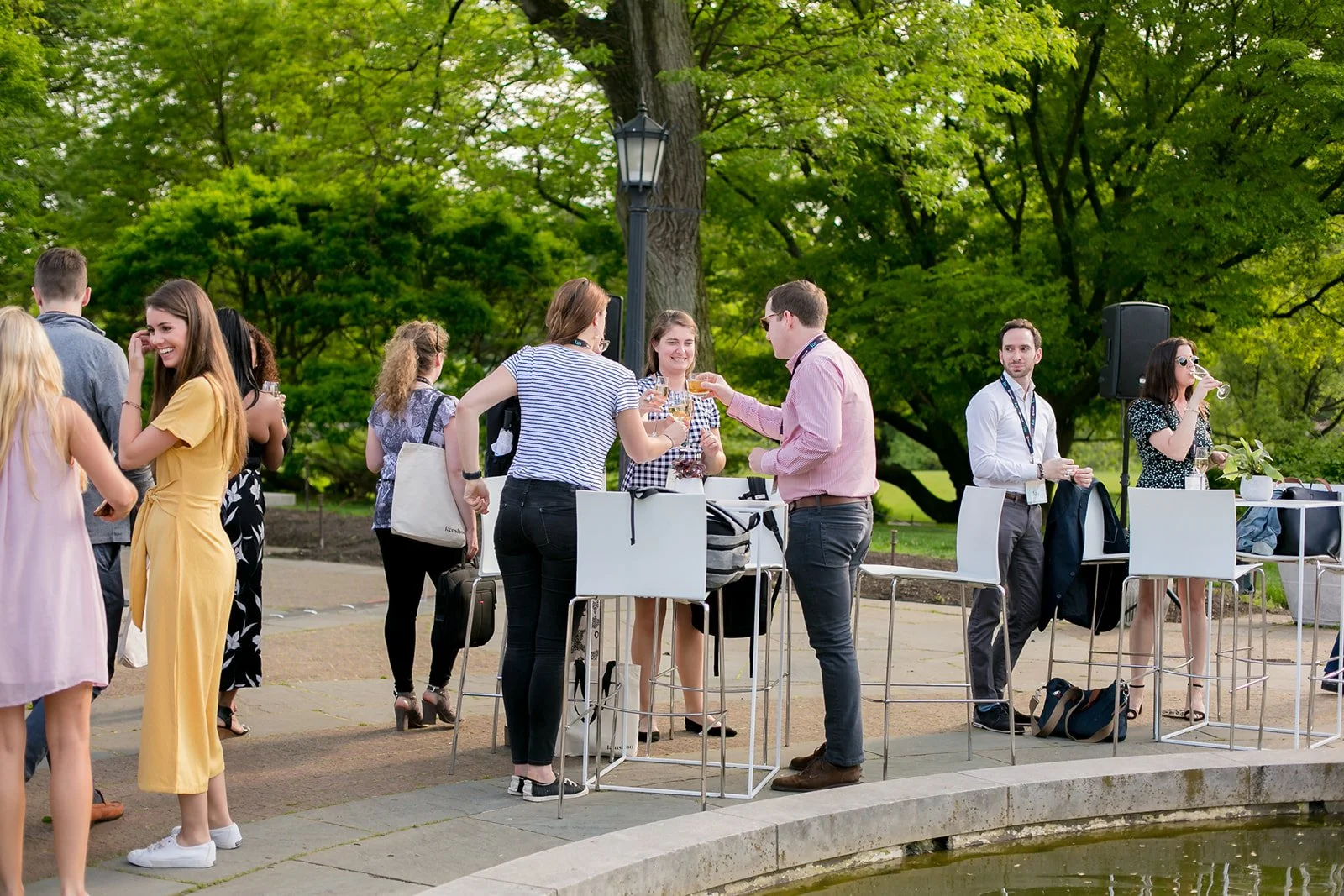 People socializing outdoors at a gathering in a park, with some standing near high tables and others around a pond, surrounded by trees.