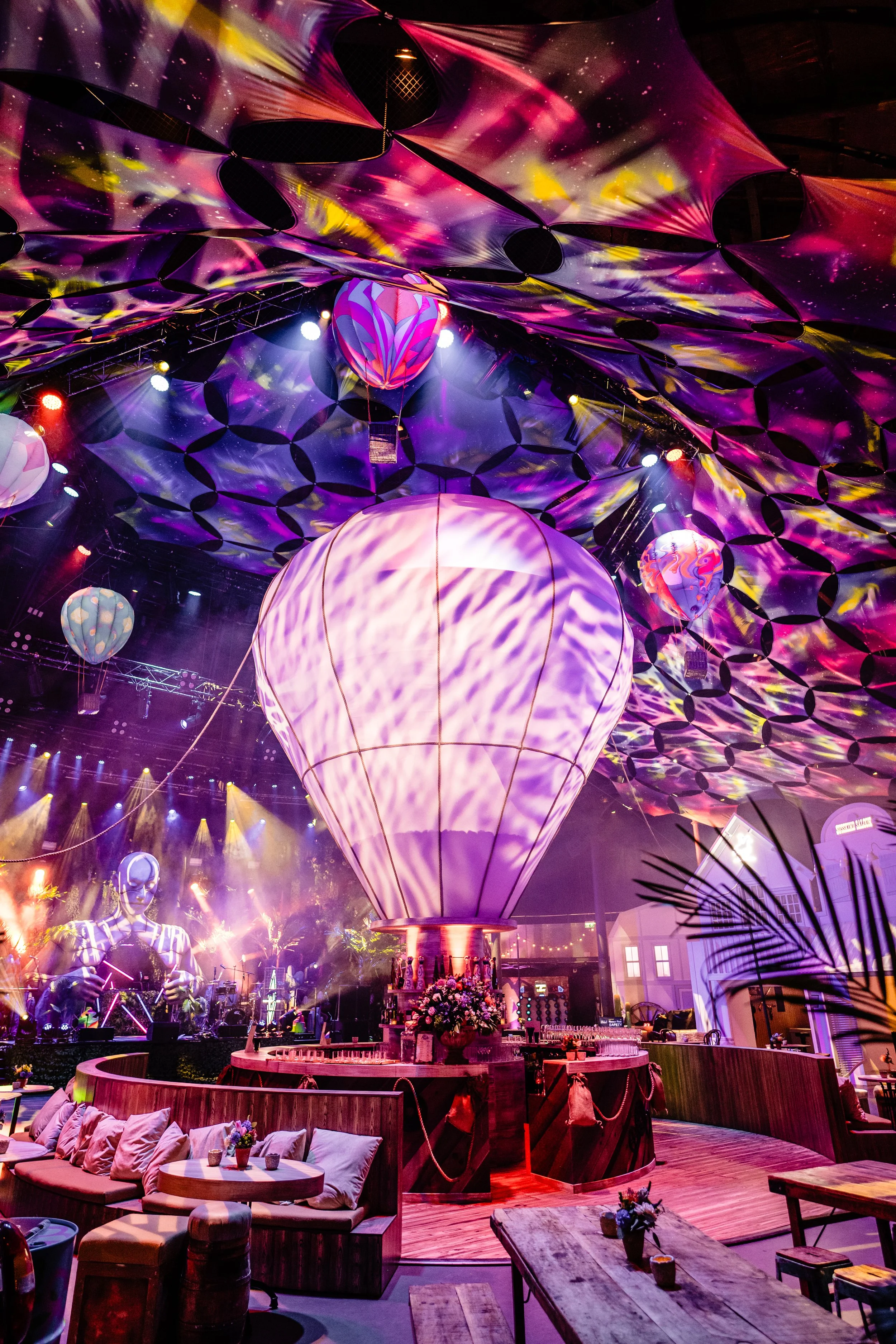 Colorful Bar Mitzvah  with large illuminated hot air balloon centerpiece, decorated ceiling with abstract patterns, hanging lanterns, stage with performer, and seating area with pillows and tables.