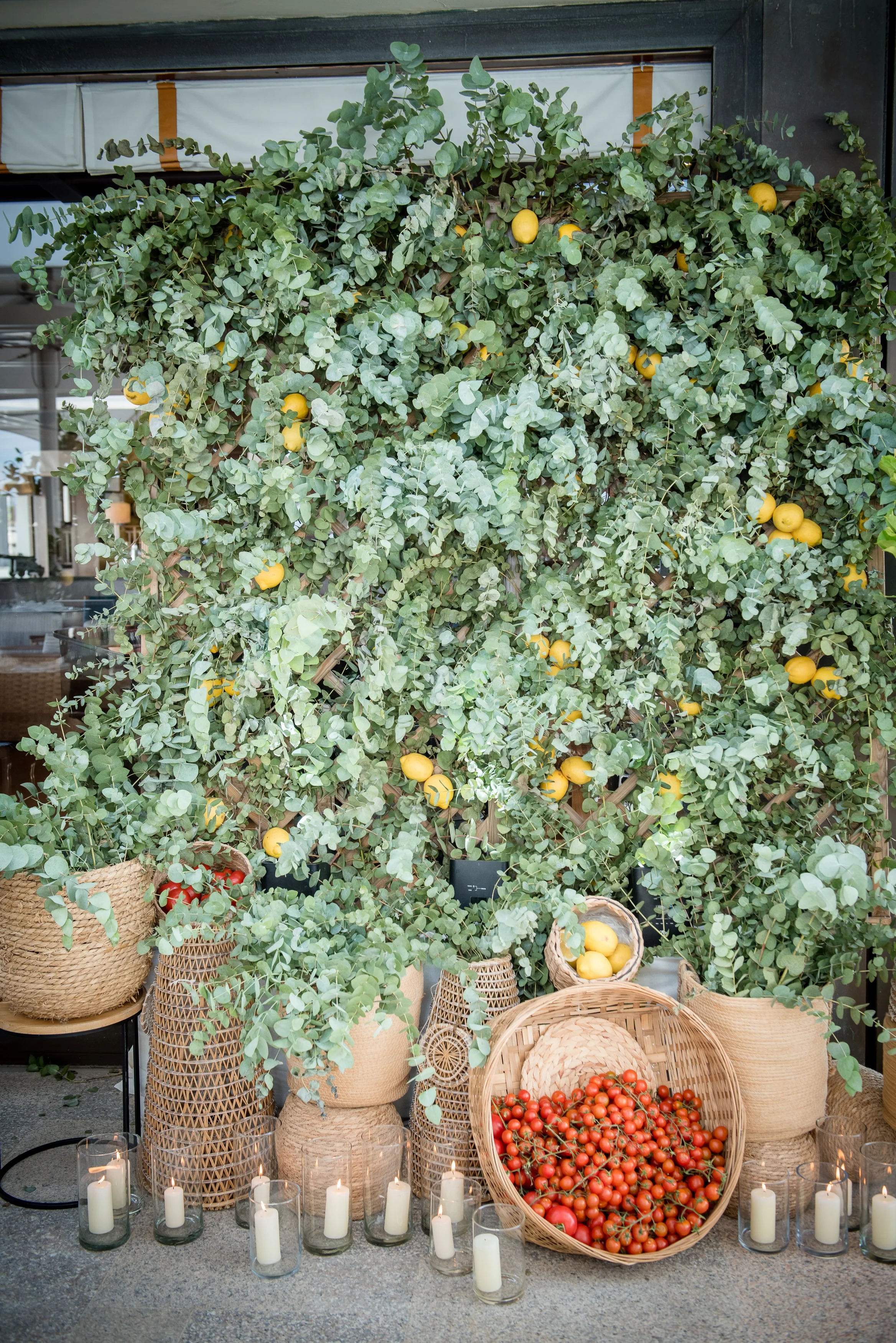 Display of fresh tomatoes and lemons in woven baskets surrounded by potted greenery, with lit candles arranged on the ground.