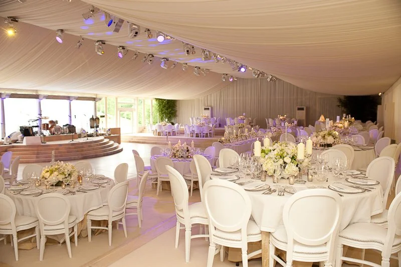 Elegant banquet hall decorated with white round tables and chairs, floral centerpieces, candles, and a small stage with a music setup under a white draped ceiling with purple and yellow lighting.