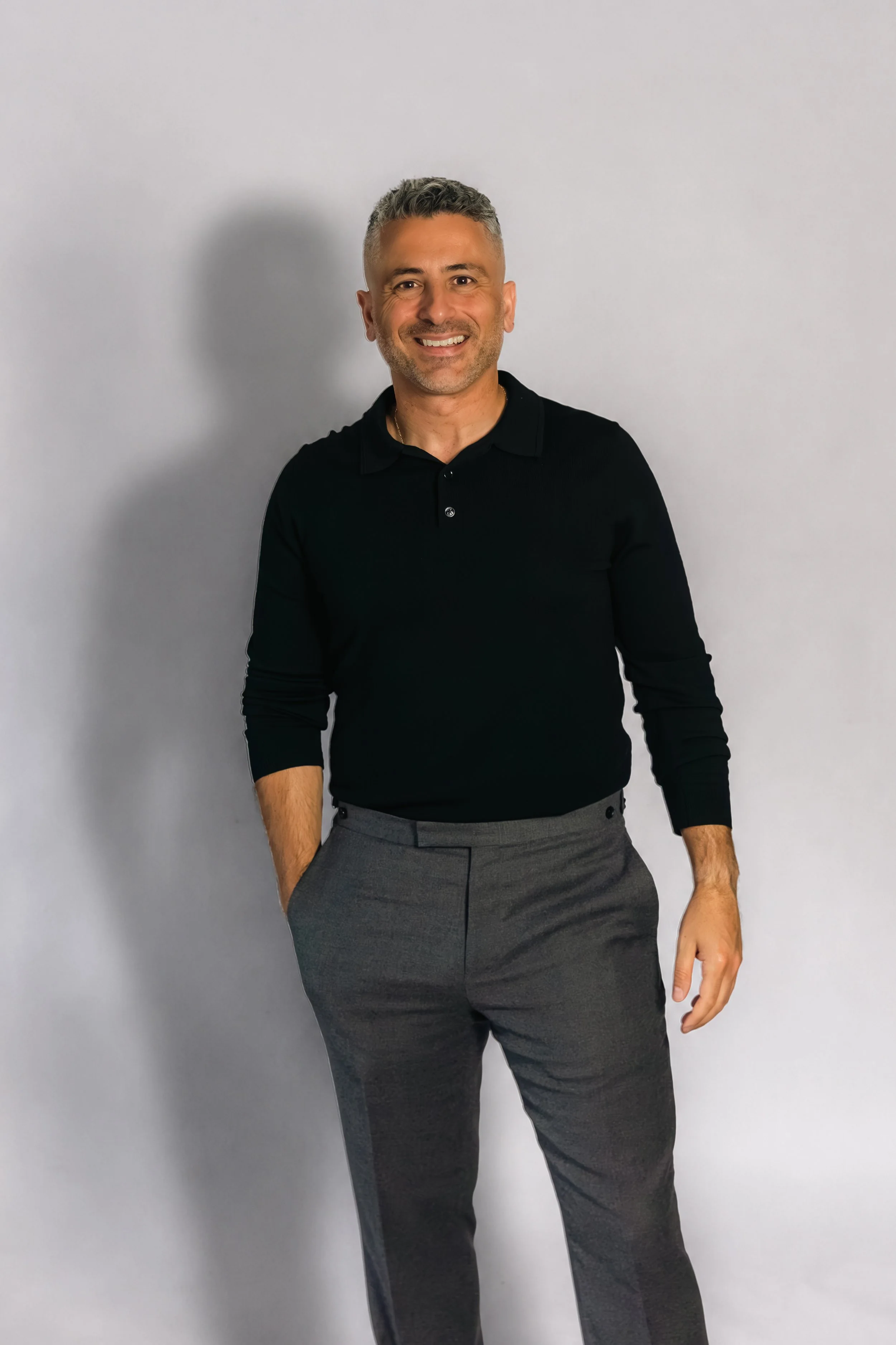 Laurie Weitzkorn and Stas Anastasiou wearing a black collared shirt and gray dress pants, standing against a plain light gray wall, smiling at the camera.