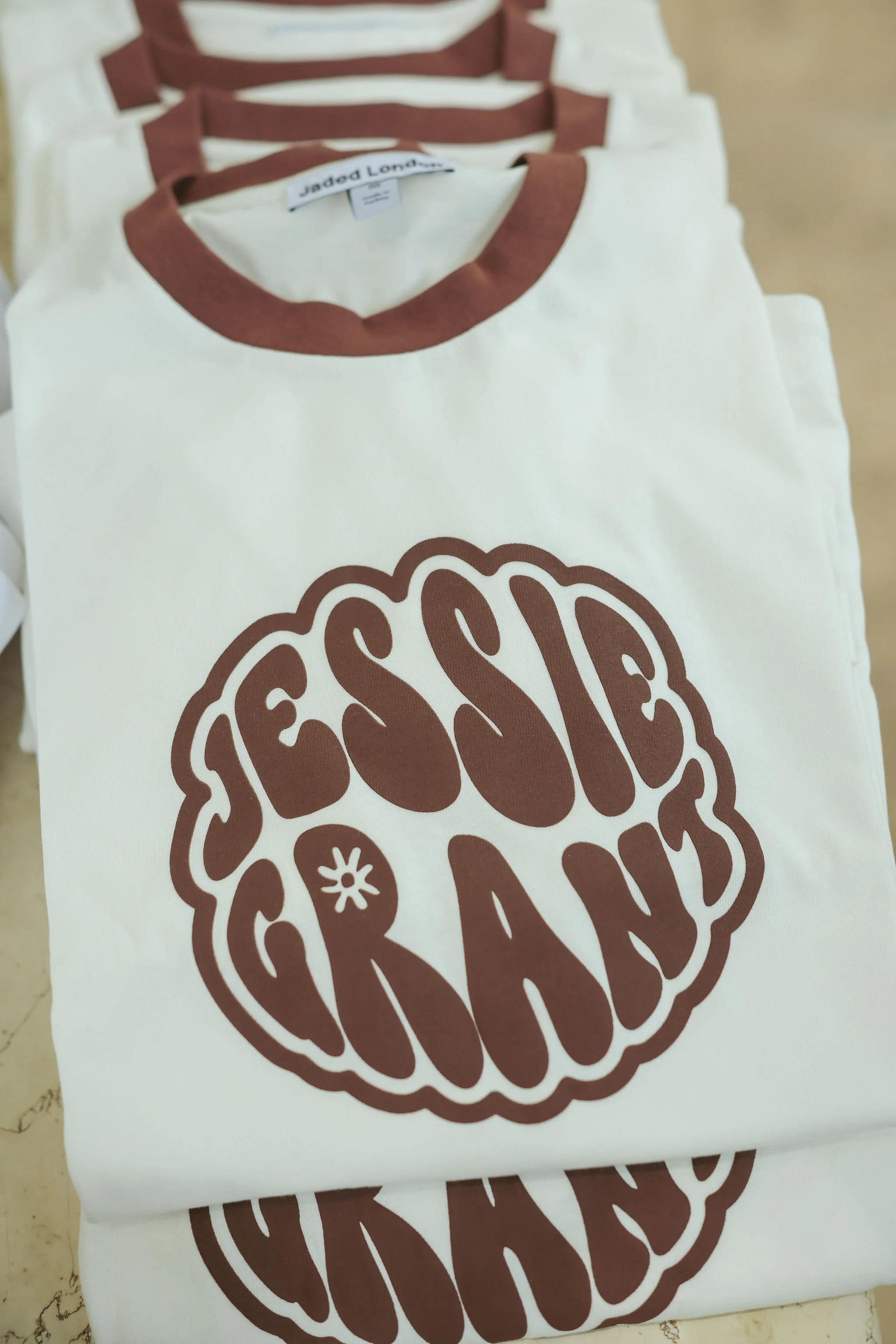 Stacked white T-shirts with brown collar and a logo of a brain with the words ' Jessie & Grant' 