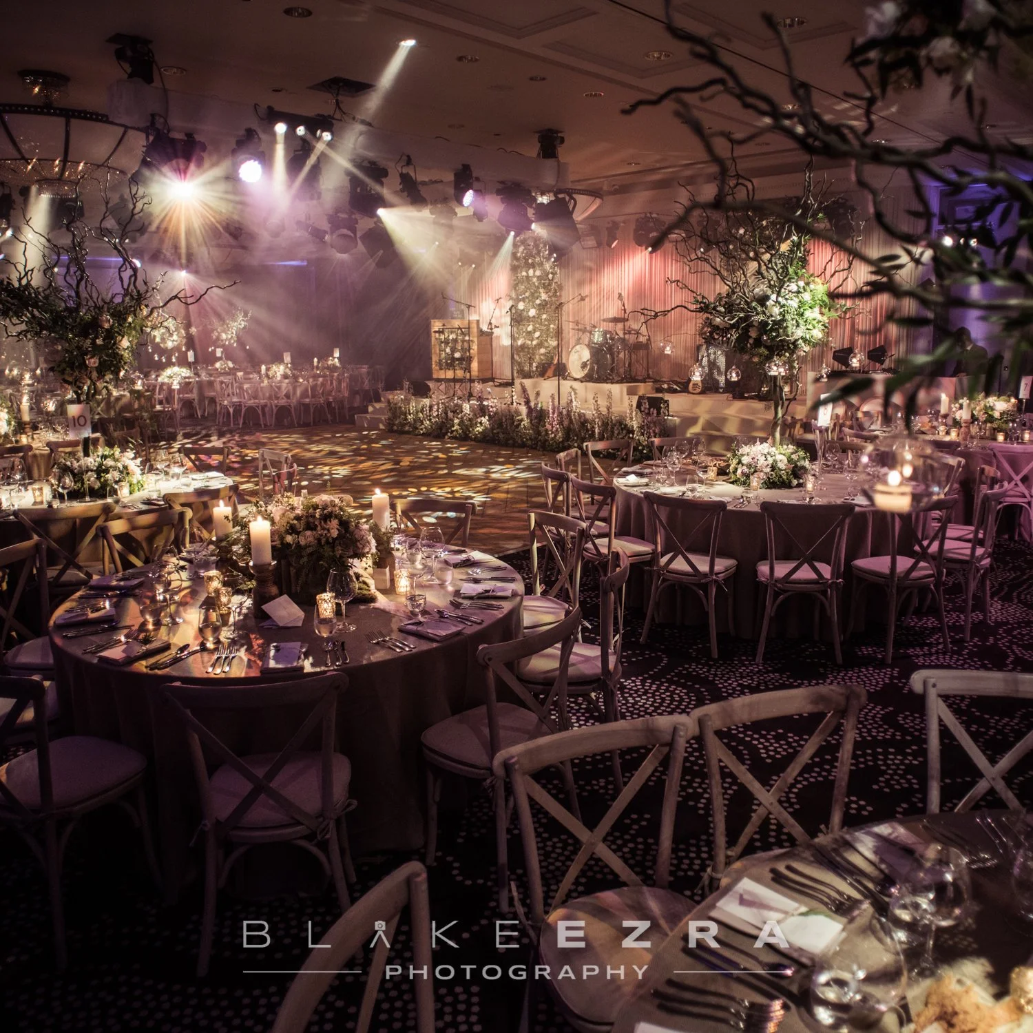 Elegant event venue decorated with floral arrangements, candles, and table settings with chairs, a stage with lights and musical instruments, and dim lighting creating a festive atmosphere. Rosewood London