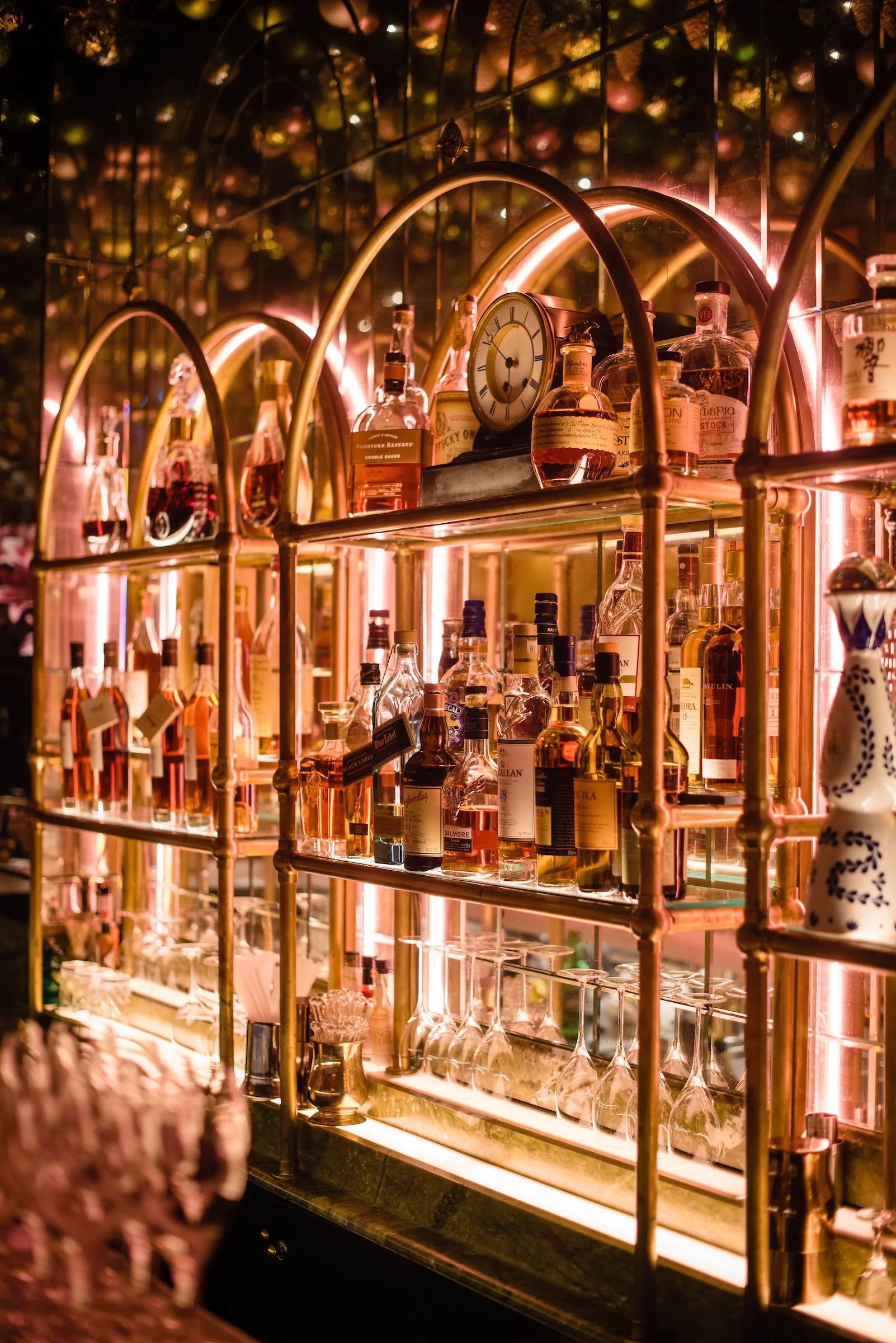 A bar shelf illuminated with warm lighting, displaying various bottles of liquor and glassware, with an ornate clock on top of the bottles.