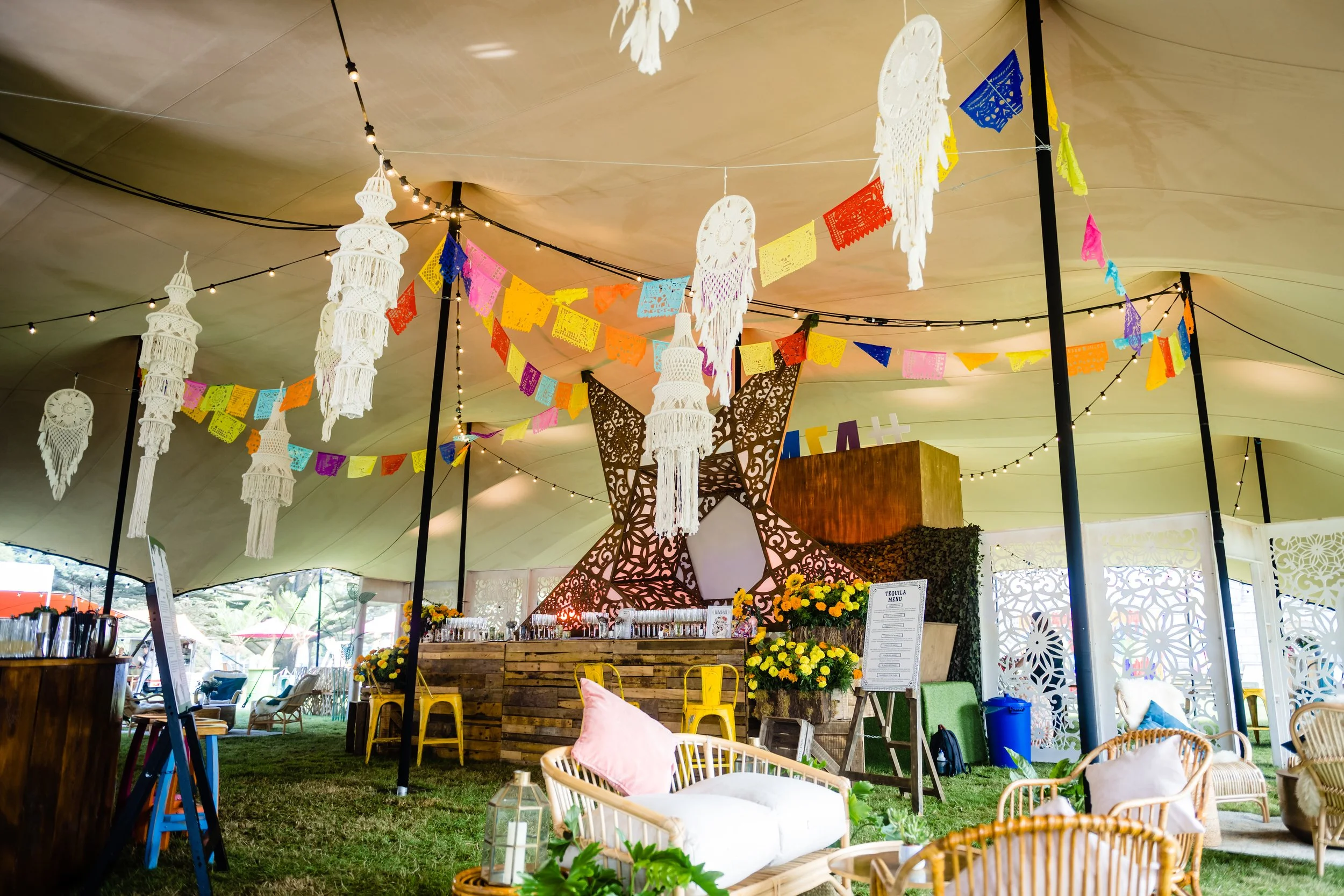 Decorated outdoor party tent with colorful papel picado banners, hanging white lace lanterns, string lights, and a variety of seating arrangements including woven chairs, cushions, and a bar area with flowers.