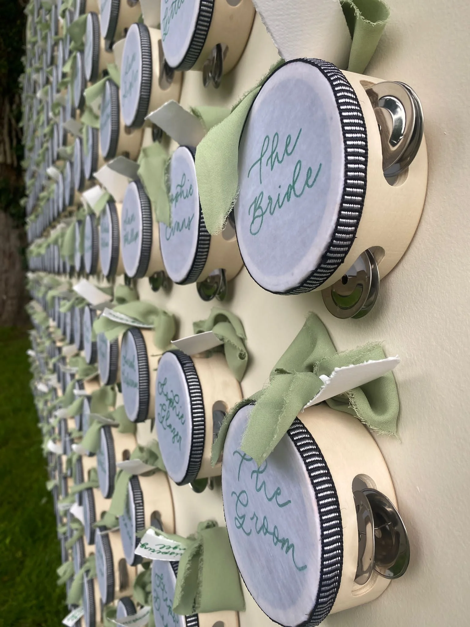 Multiple small jars with black and white striped lids lined up on a table, each with a piece of light green fabric and a white paper beneath the lid that has handwritten names in blue ink, such as 'The Bricks'.