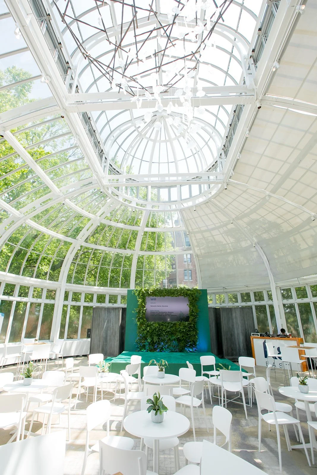 Inside a bright, modern greenhouse with a glass ceiling and walls, white chairs and tables with small potted plants, and a green backdrop with a screen, in an event space.