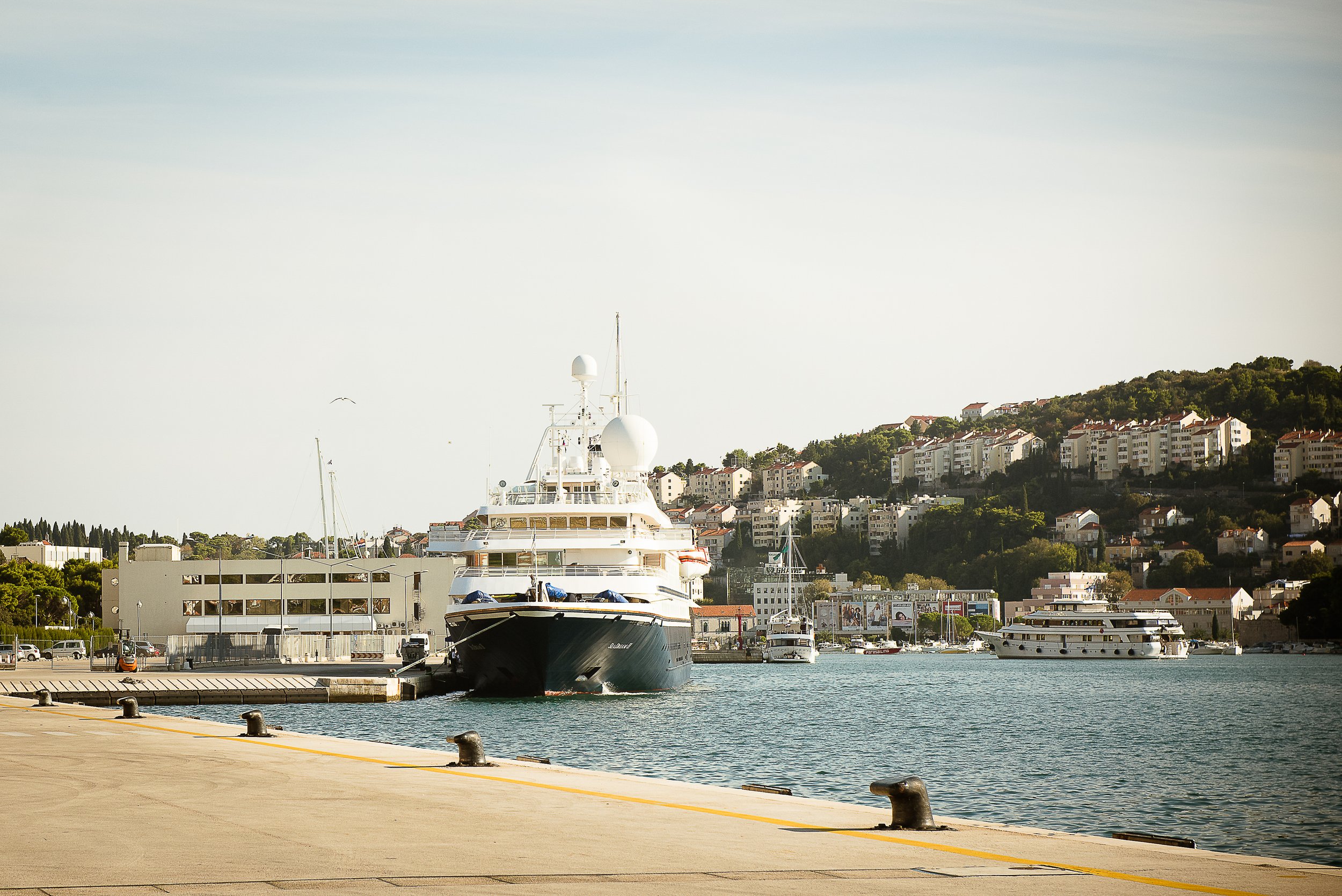 Large yacht docked at marina with buildings and a hillside in the background.