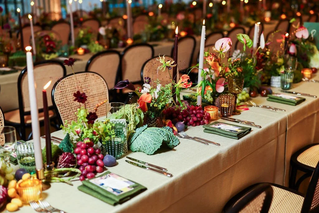 A long banquet table decorated with a floral arrangement featuring fruits, vegetables, and flowers, lit candles, and place settings with napkins and cutlery, set for a wedding dinner.
